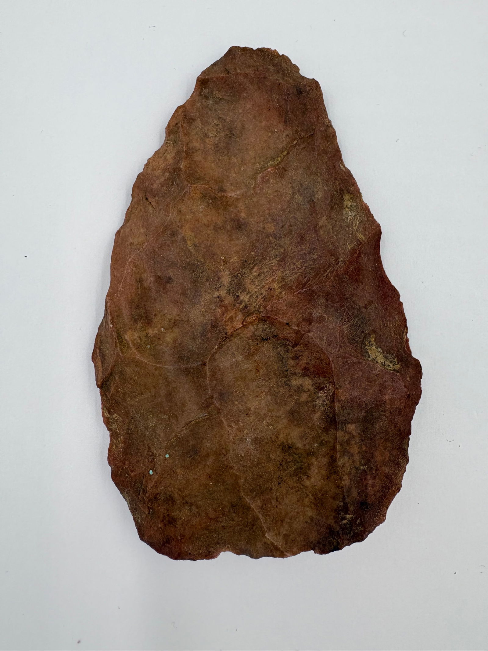 North Blade: Flaked reddish-brown stone blade with a teardrop profile and bifacial working. Surface displays prominent flake scars, subtle mineral streaking, and irregularly retouched edges leading to a narrow