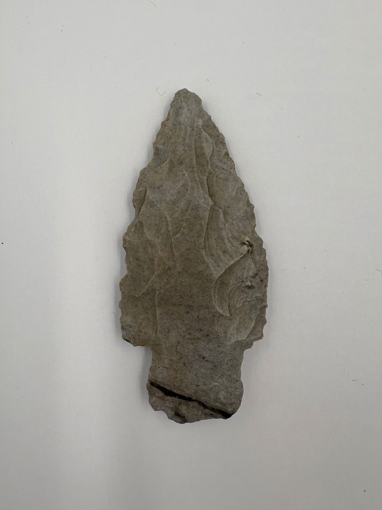 Adena: Flaked stone projectile point measuring 3in with a lanceolate outline and narrow, tapered tip. Bifacial flake removal along both faces forms a thinned blade with irregular lateral notches. Gray-brown