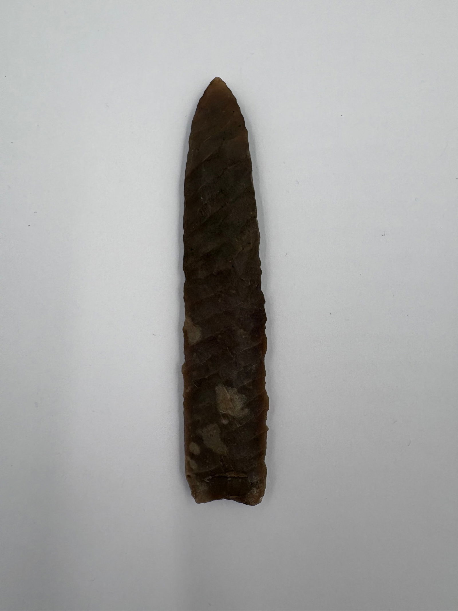 *Restored* Eden: Long, narrow flaked stone blade measuring 4in in length. Rich brown tones with subtle lighter mottling and pronounced bifacial flake scars along the faces, producing a ridged texture. Tapered, acute