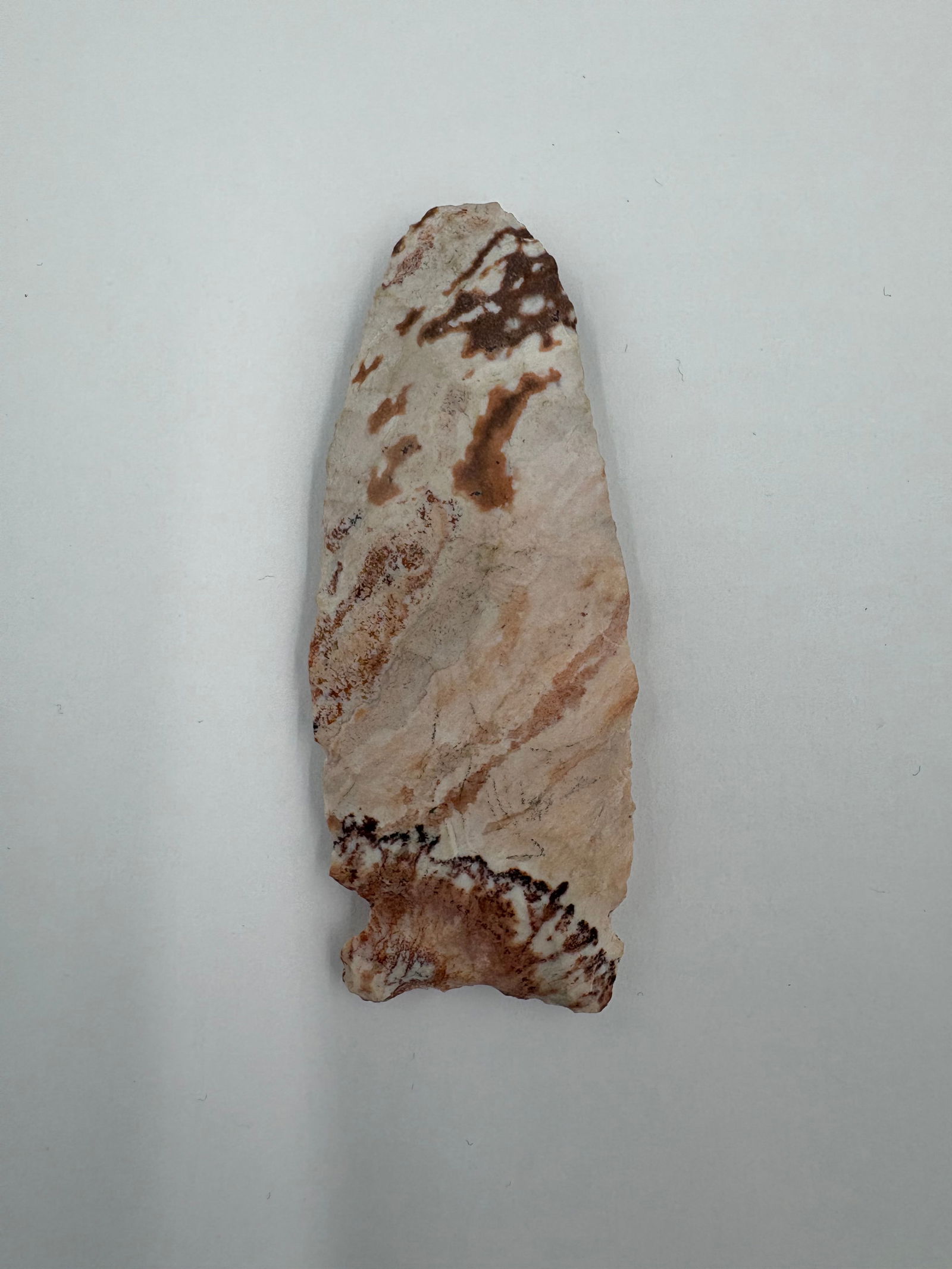 Osceola, Missouri creek find: Elongated flaked stone point measuring 3in with a lanceolate profile and tapered tip. Creamy to pale-pink ground with rust-brown and ochre mineral banding and dendritic staining across the faces.
