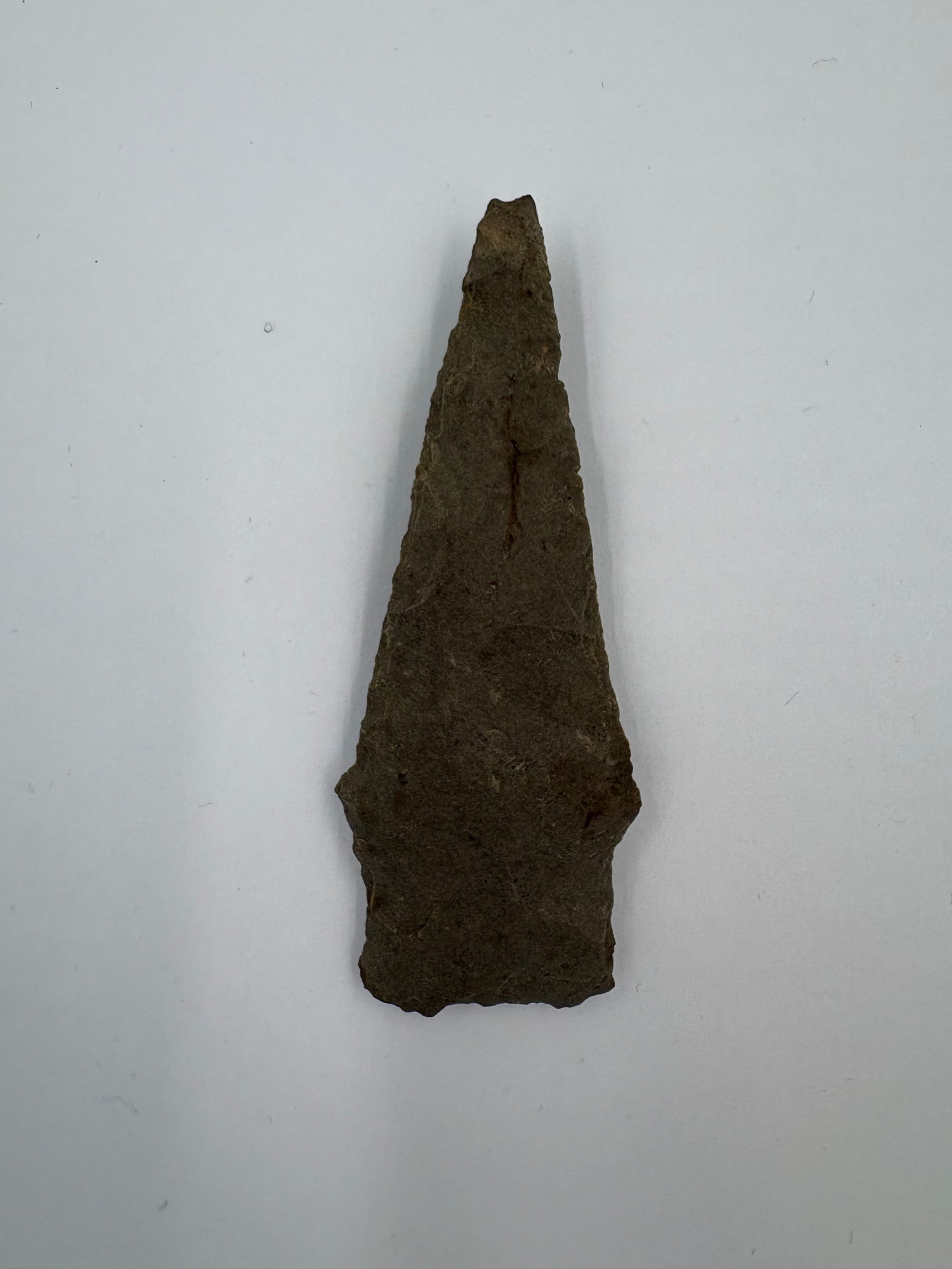 Kramer: 3 1/4-inch dark brown flaked stone point with a narrow, tapered triangular blade and subtle barbed shoulders leading to a straight stemmed base. Flake scars and a rough, weathered surface texture run
