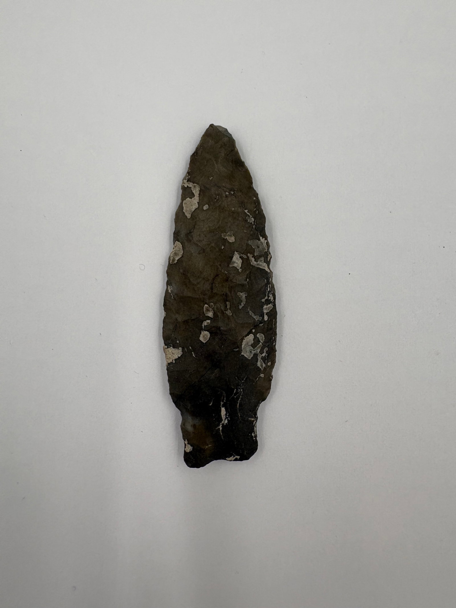 Kramer: Leaf-shaped flaked stone point measuring 2 3/4in. Dark gray to brown chert displays bifacial flake scars and a slightly contracting basal stem. Surface shows orange-brown staining and areas of white m