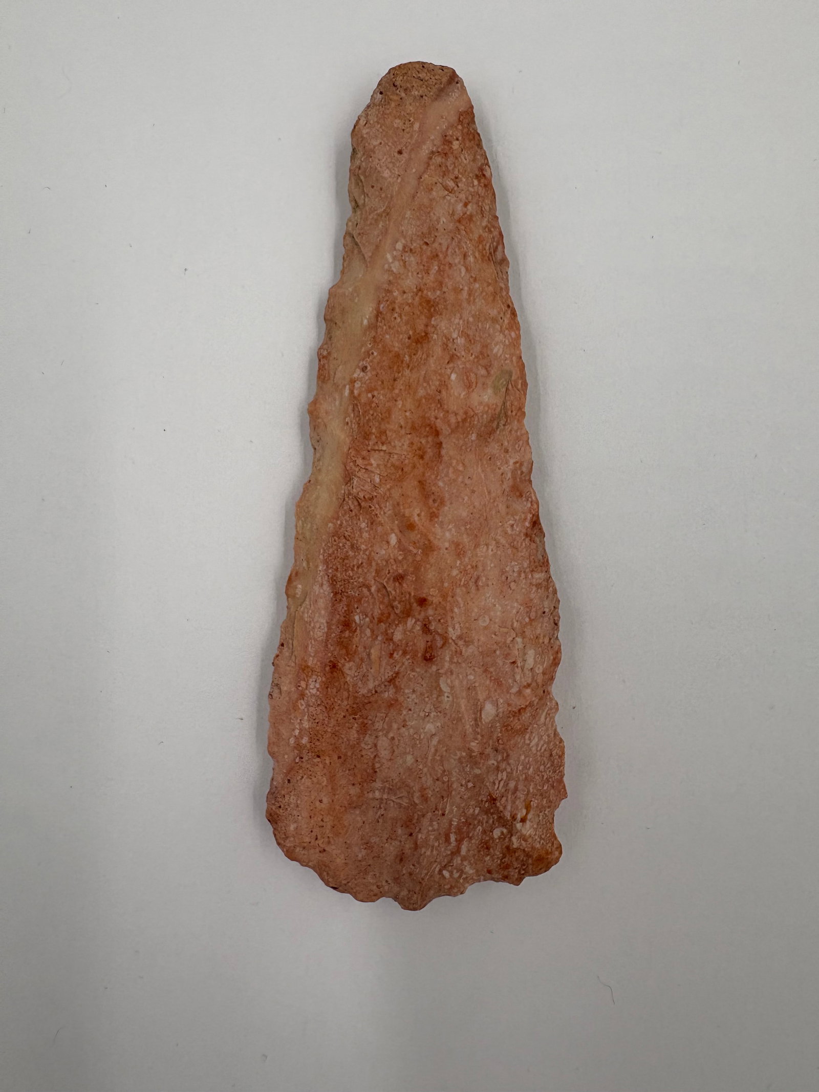 Carter Blade: Flaked stone blade measuring 4 1/2 in long, with a tapered triangular form and a broader, slightly scalloped base. Warm pink to salmon coloration with subtle lighter banding and a granular stone