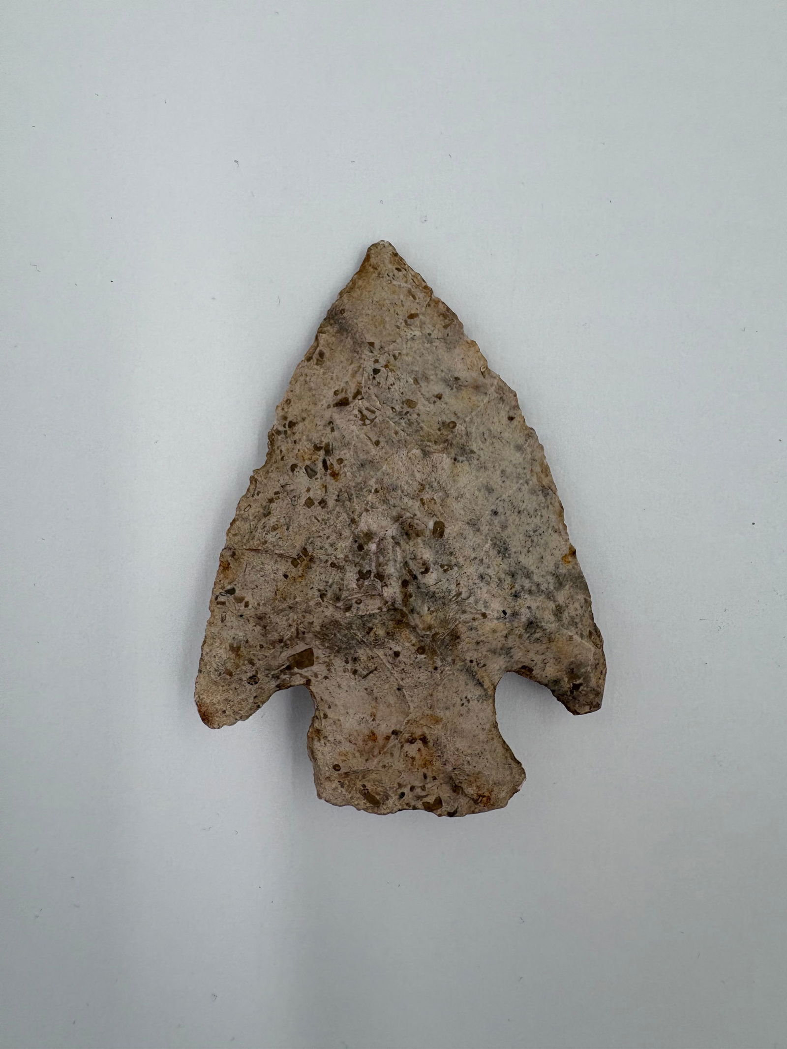 Snyder: Stone projectile point measuring 2 3/4 in with a triangular blade and a short stemmed base featuring pronounced side notches. Beige-gray surface shows brown mineral flecks and a coarse texture from bi