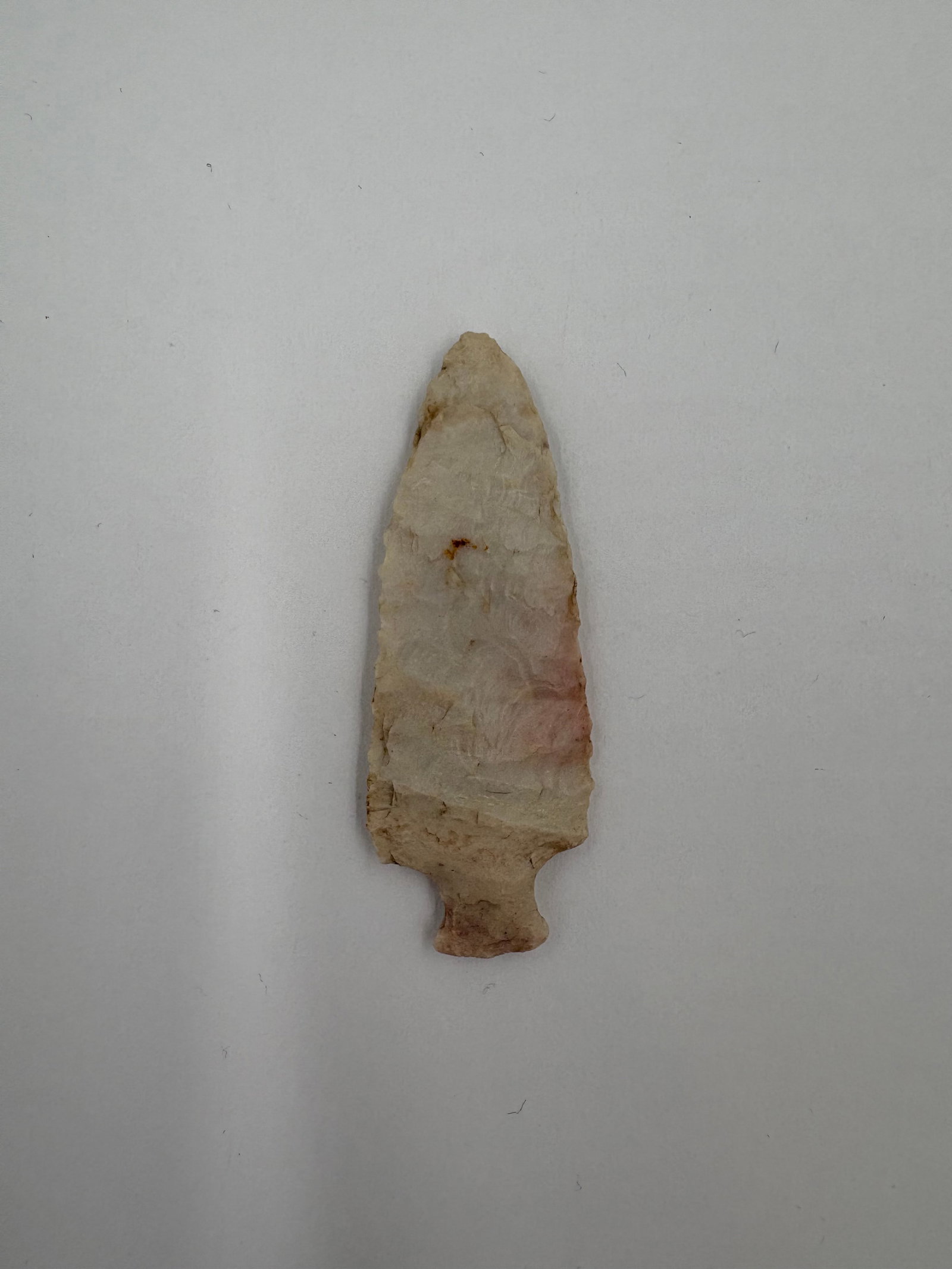 Table Rock: Light tan to pale pink flaked stone point with a narrow stem, expanding shoulders and a tapered triangular blade. Bifacial flake scars cover both faces and the slightly concave base leads into a