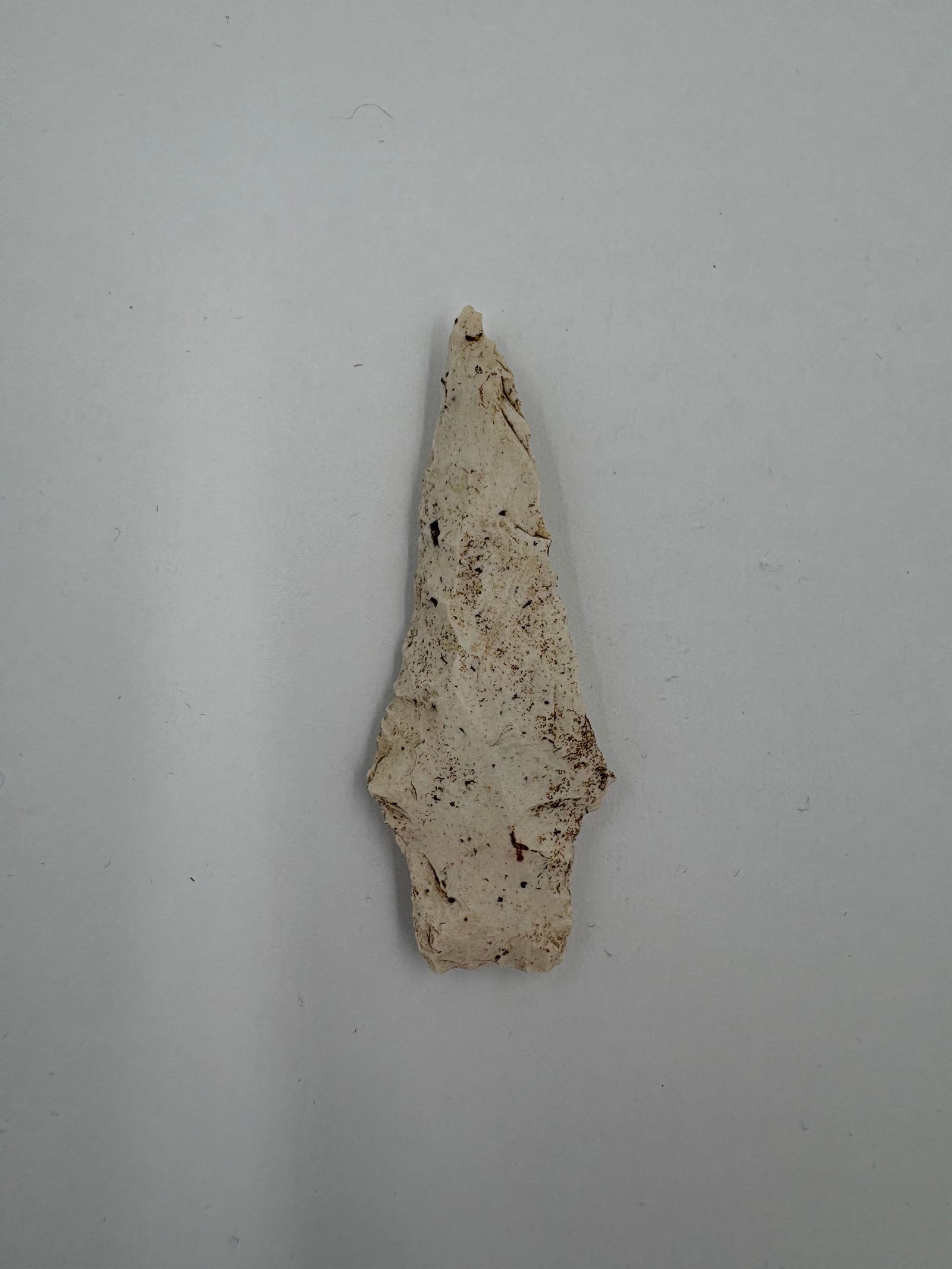 Dickson: Elongated cream-colored flaked stone point with a narrow triangular blade, slight shoulders and a defined stemmed base measuring 2 5/8 in. Bifacial flake scars create a textured surface with brown