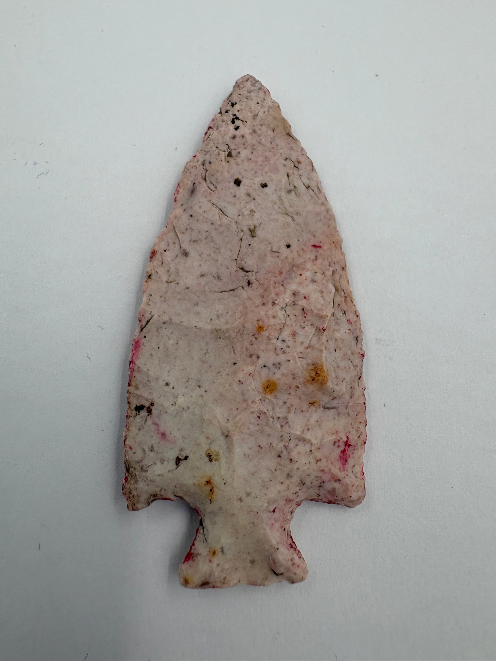 Kay Blade: 2 3/8 in stemmed projectile point with a triangular blade and a notched, flaring basal stem. Light cream to pale pink surface with red and yellow flecks and scattered dark mineral inclusions. Thin,
