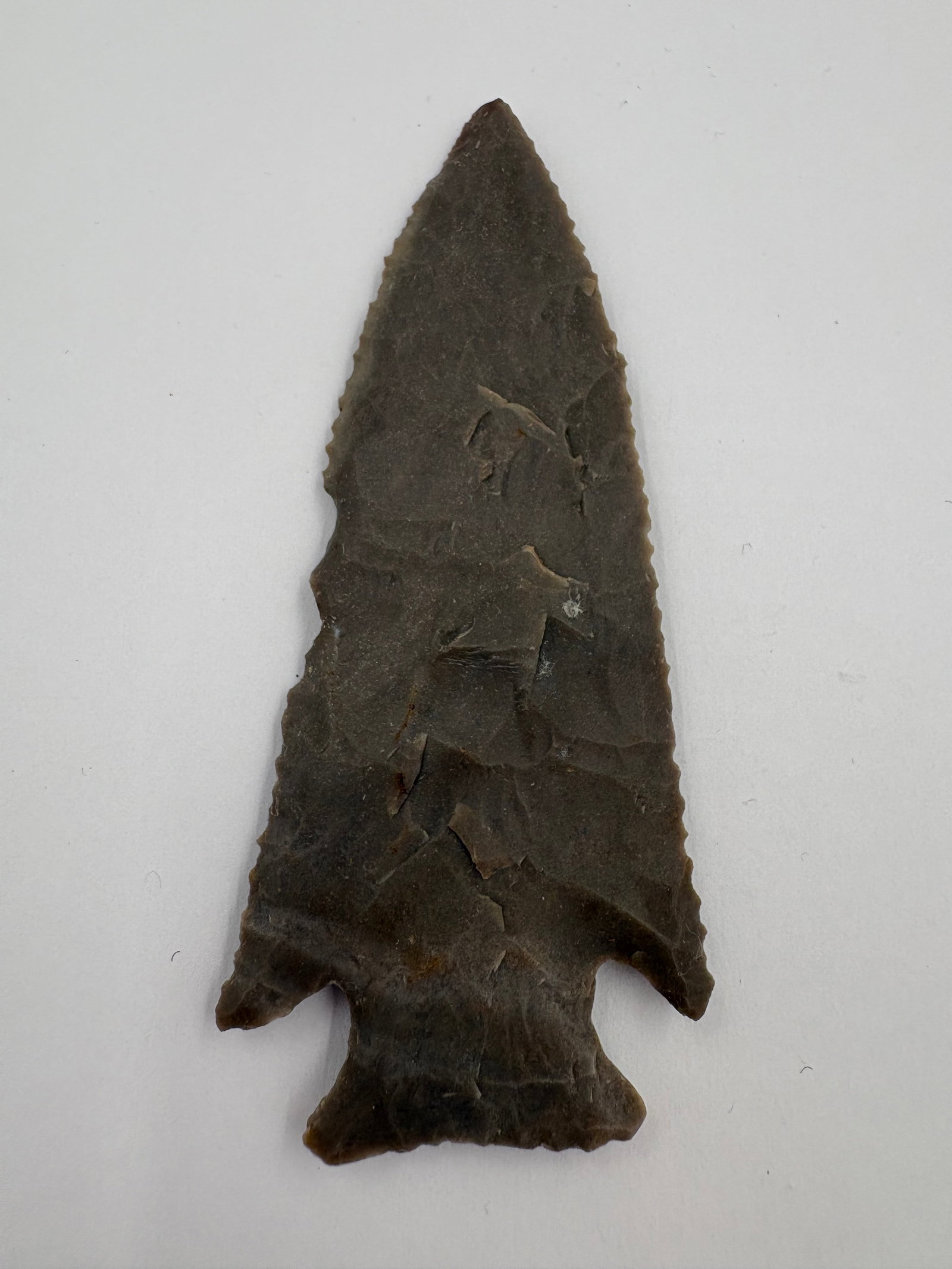 Hardin: Hardin-style stone projectile point measures 3 7/8in with a slim, triangular blade that tapers to a sharp tip. Green-gray to dark brown material displays bifacial flaking and fine serration along the