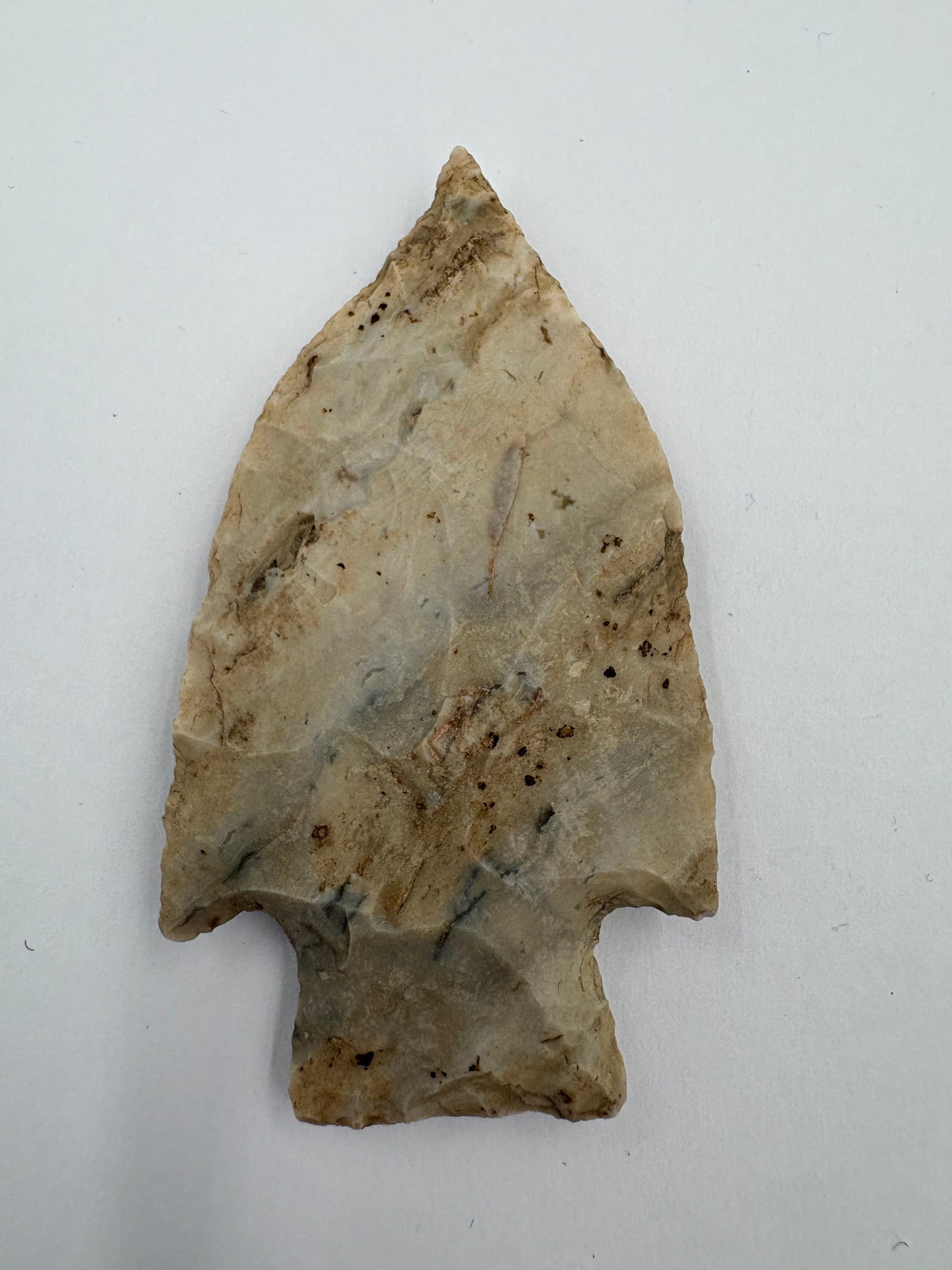 Etley: Flaked chert projectile point measuring 2 3/4 in with a triangular blade, concave shoulders and a short stemmed base. Pale beige to gray coloration with darker mineral streaks and scattered brown