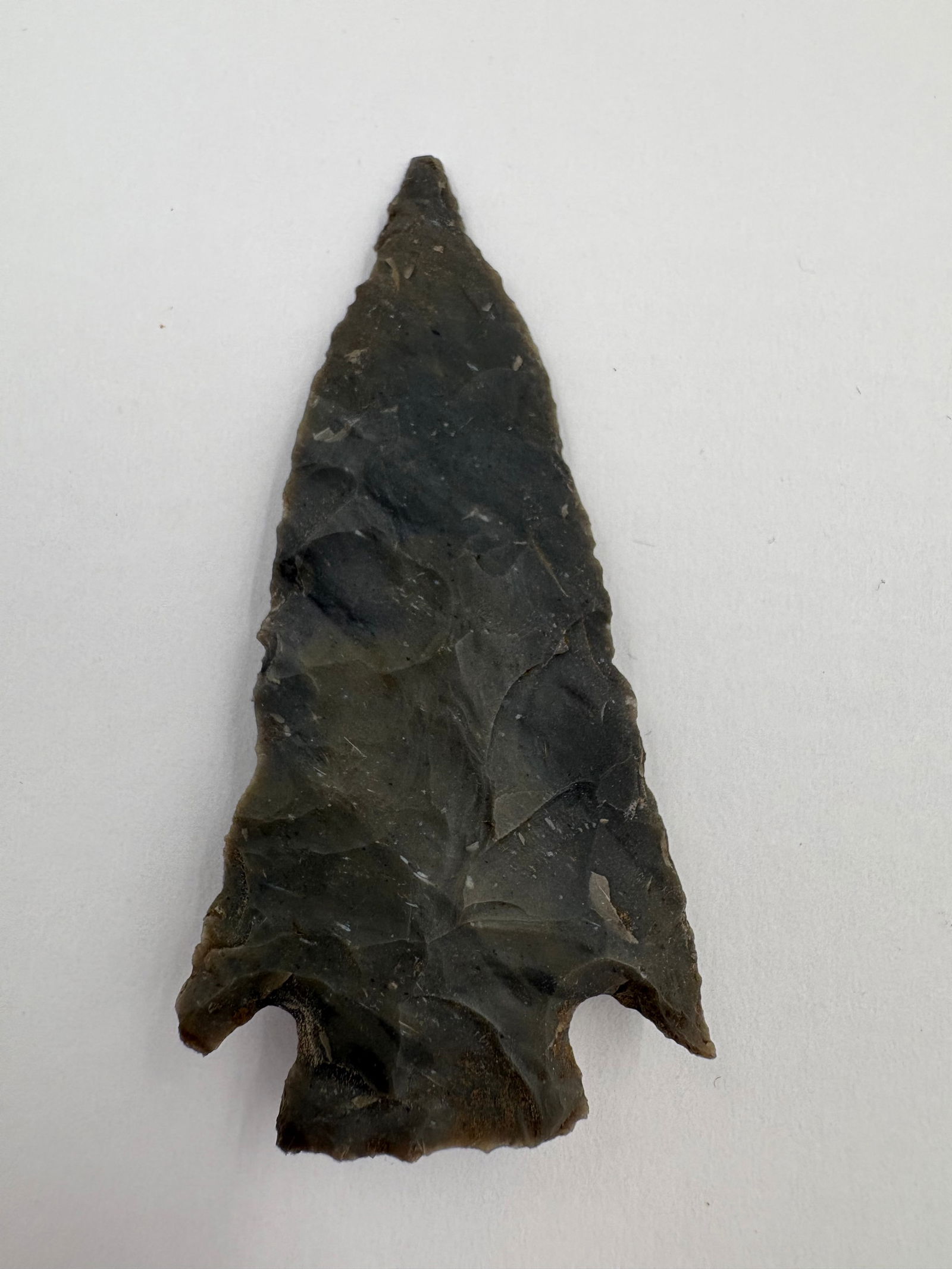 Pine Tree: Dark gray to black hand-knapped chert projectile point with a broad triangular blade, narrow stem and pronounced basal notches. Flake scars and pressure-retouch textures cover both faces, and an