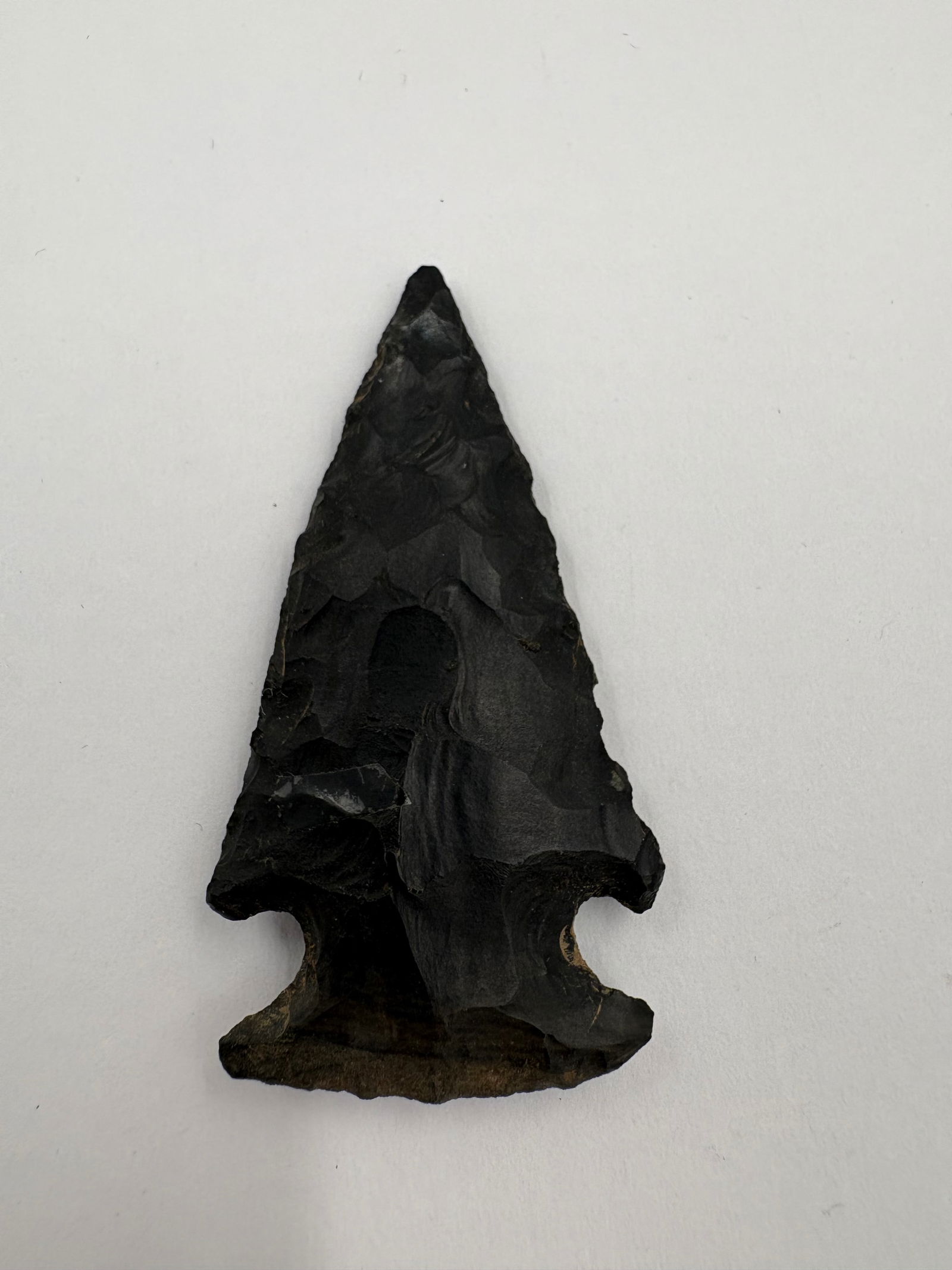 Hopewell: Black, bifacially flaked stone projectile point with pronounced side notches and a broad stemmed base. Distinct flake scars cover both faces and the narrow triangular blade terminates in a