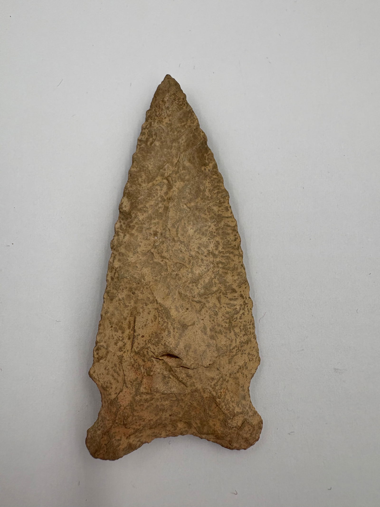Hemphill: Leaf-shaped flaked stone projectile point measuring 2 3/4in. Light tan to beige stone with bifacial flaking and serrated edge retouch along the blade. Distinct stemmed base with prominent basal ears a