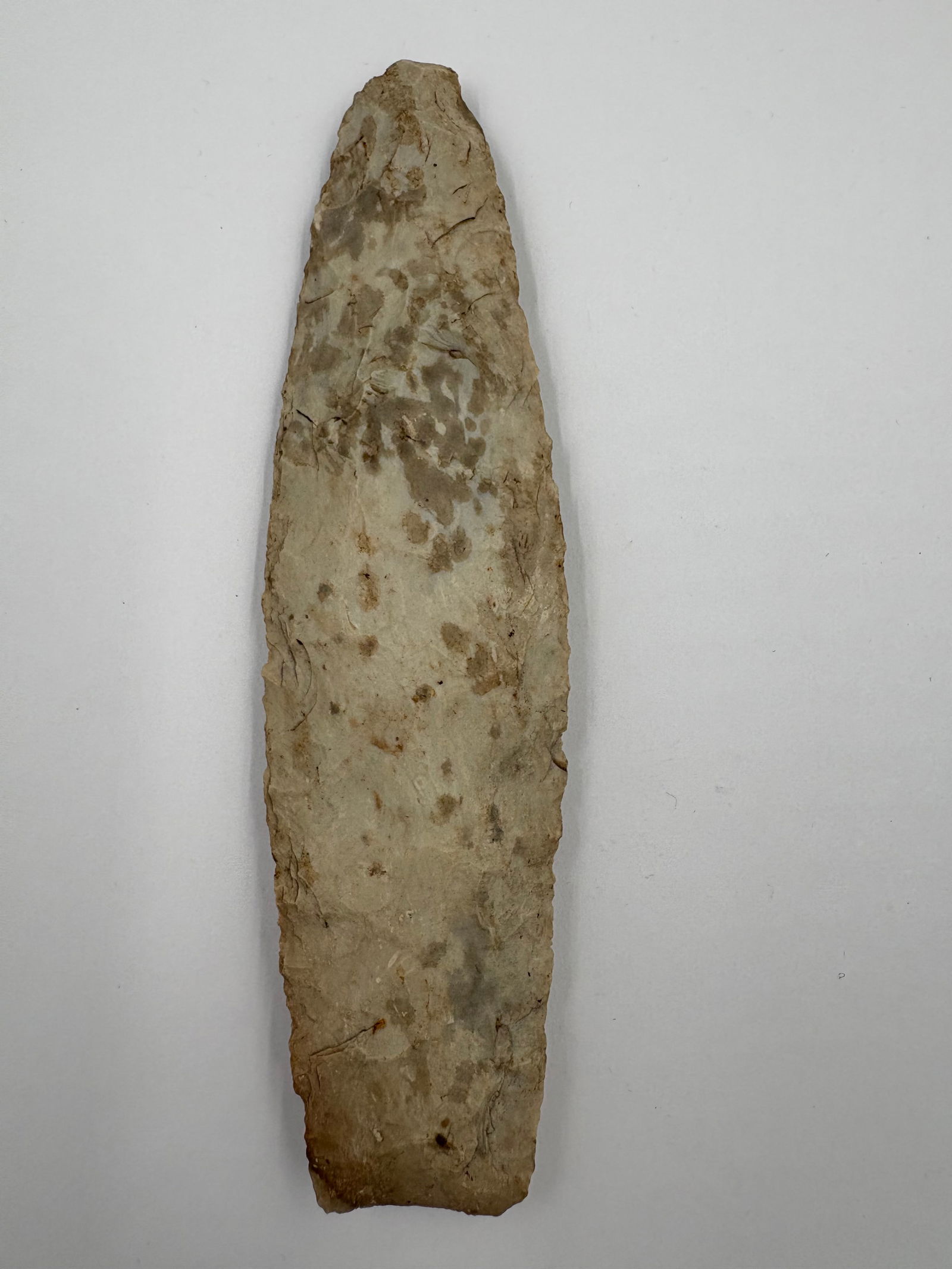 Sedalia: Long, narrow flaked stone blade with an elongated lanceolate outline, a tapered tip and a broader squared base. Light tan to gray coloration with bifacial flake scars, edge retouch and small chips