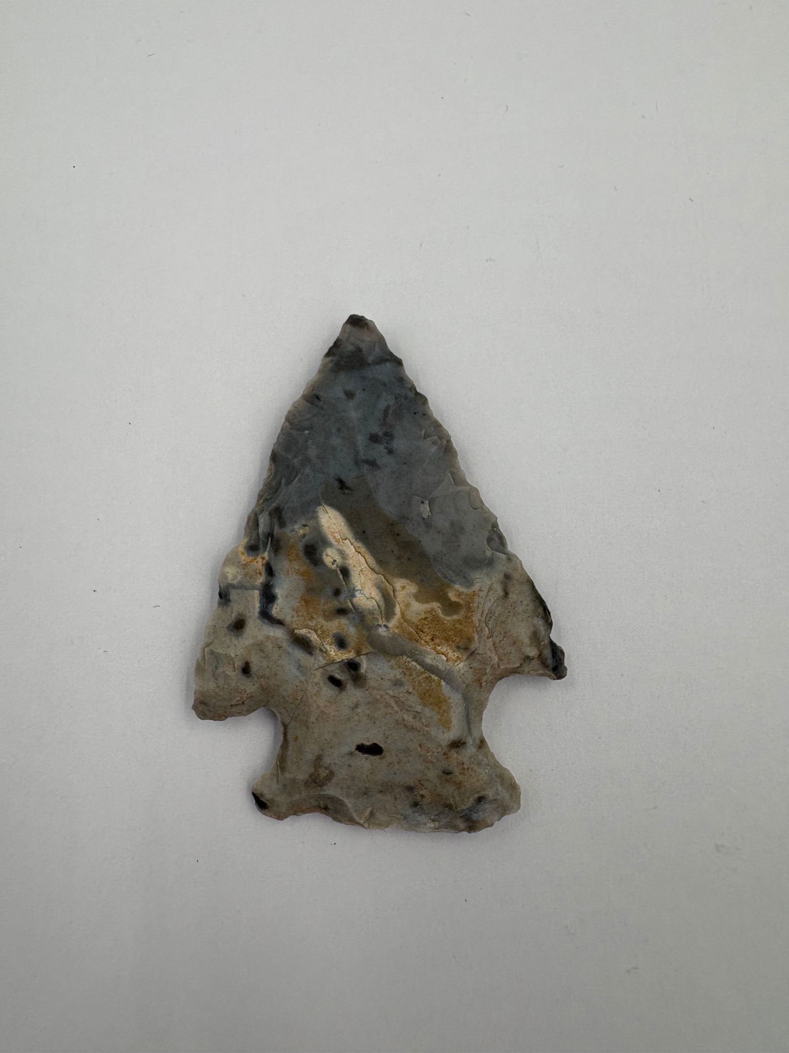 Snyder: Chert projectile point with a triangular blade, pronounced notched base and flared basal ears, exhibiting bifacial knapping and distinct flake scars. Tan and brown mineral staining contrasts the gray