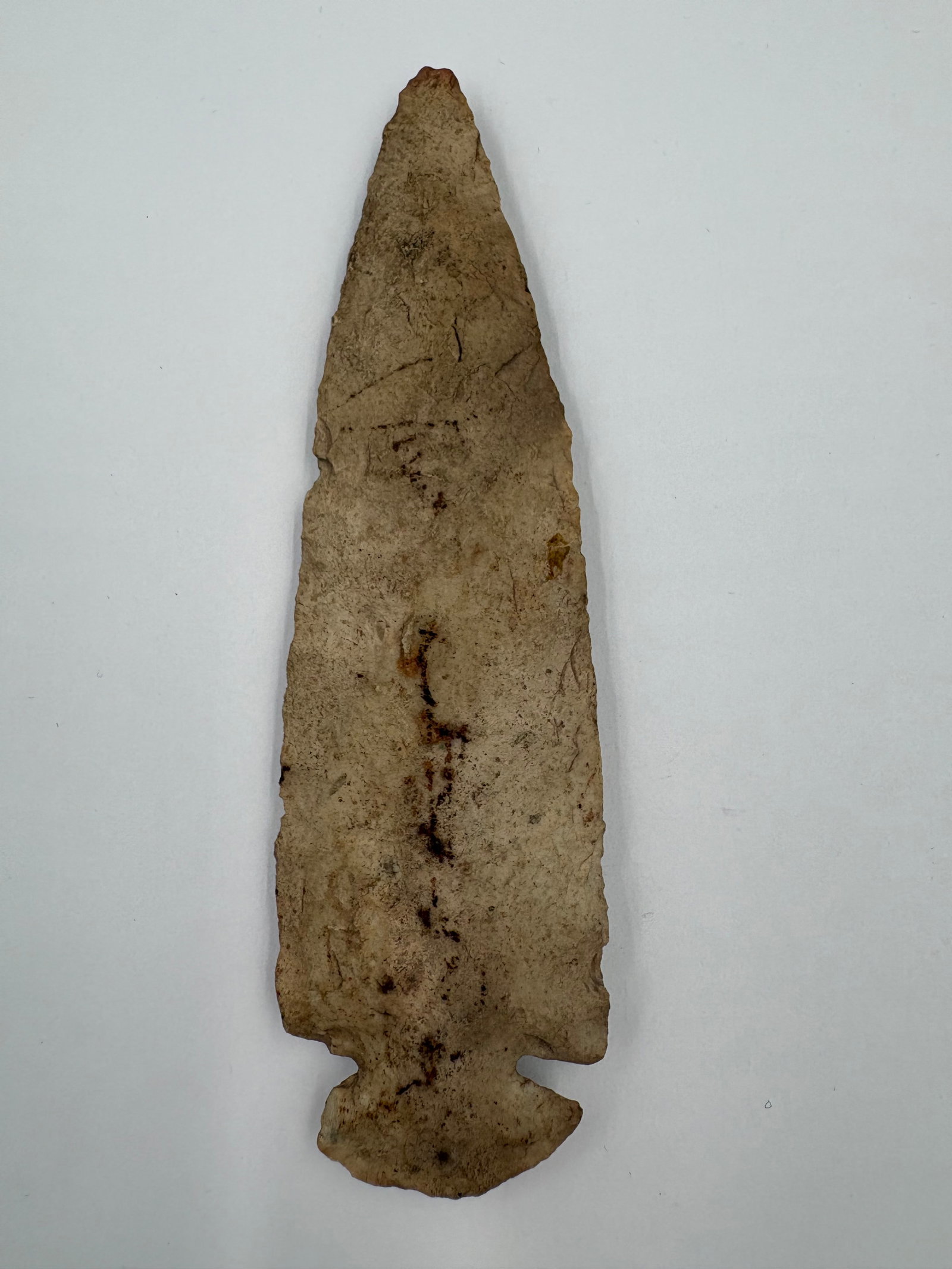 Dovetail: Hand-flaked stone projectile point with a long, narrow triangular blade and a notched, stemmed base. Pale beige to tan coloration with brown and rust-colored staining concentrated along the