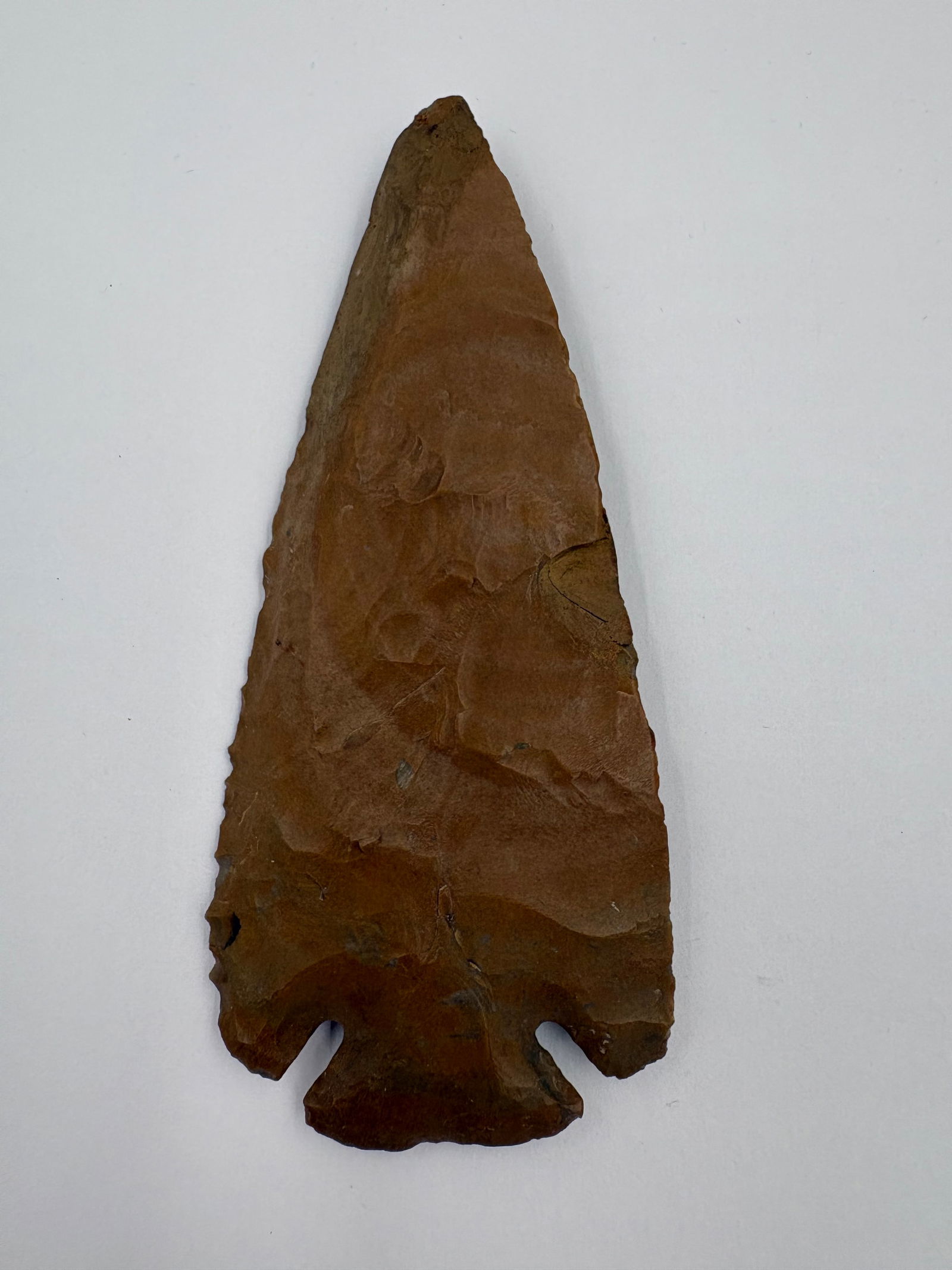 Dovetail (minor restoration): Brown flaked stone projectile point with a pronounced dovetail base and paired basal notches. Measures 4 1/4in long and displays bifacial flaking with pressure-flake scars across the blade and a