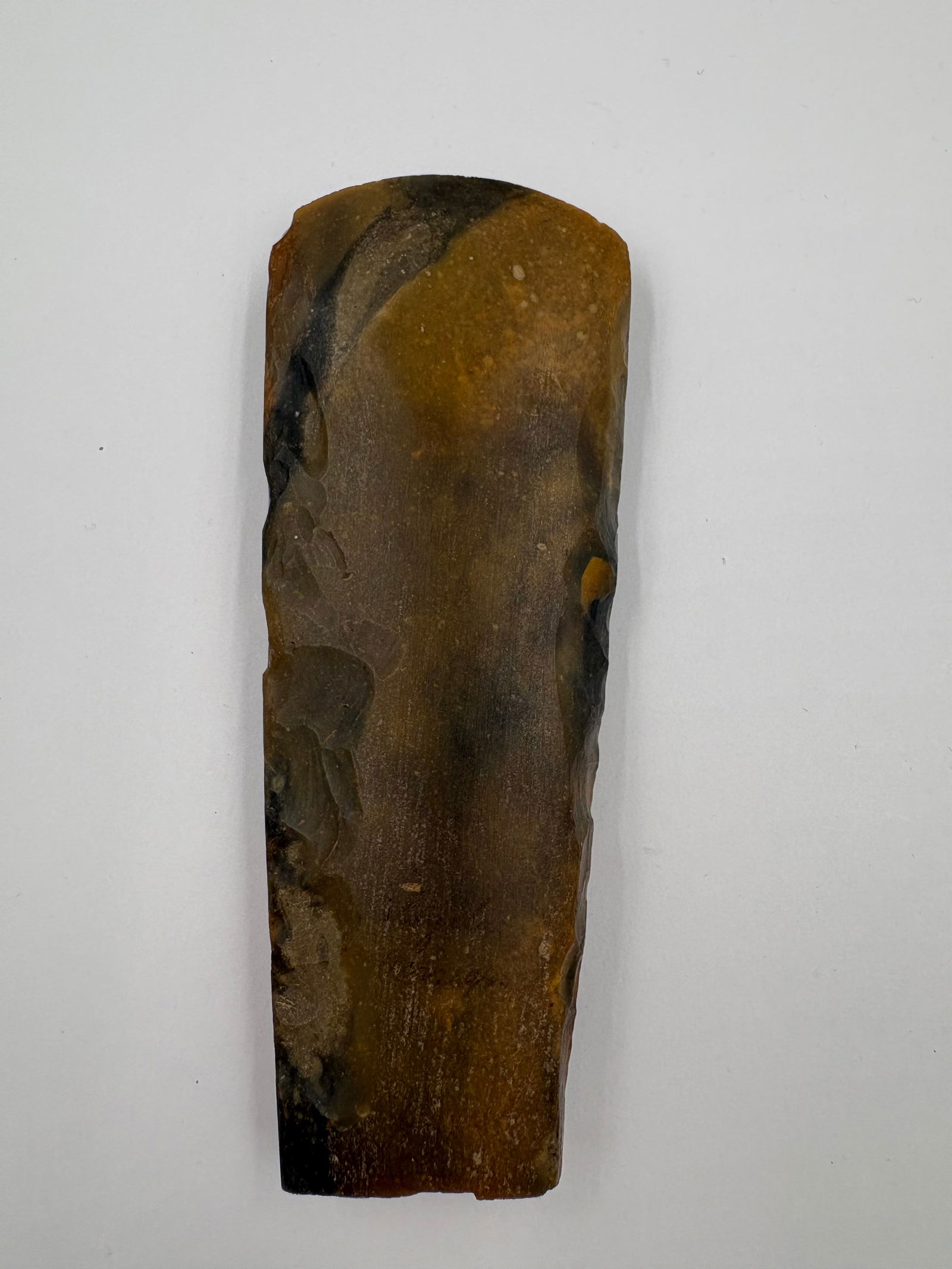 Danish Celt: Hand-flaked stone celt measuring 5 1/4 in long, with a tapered, slightly pointed end and a squared base. Broad flake scars and localized smoothing run along the faces, with evidence of edge retouch.