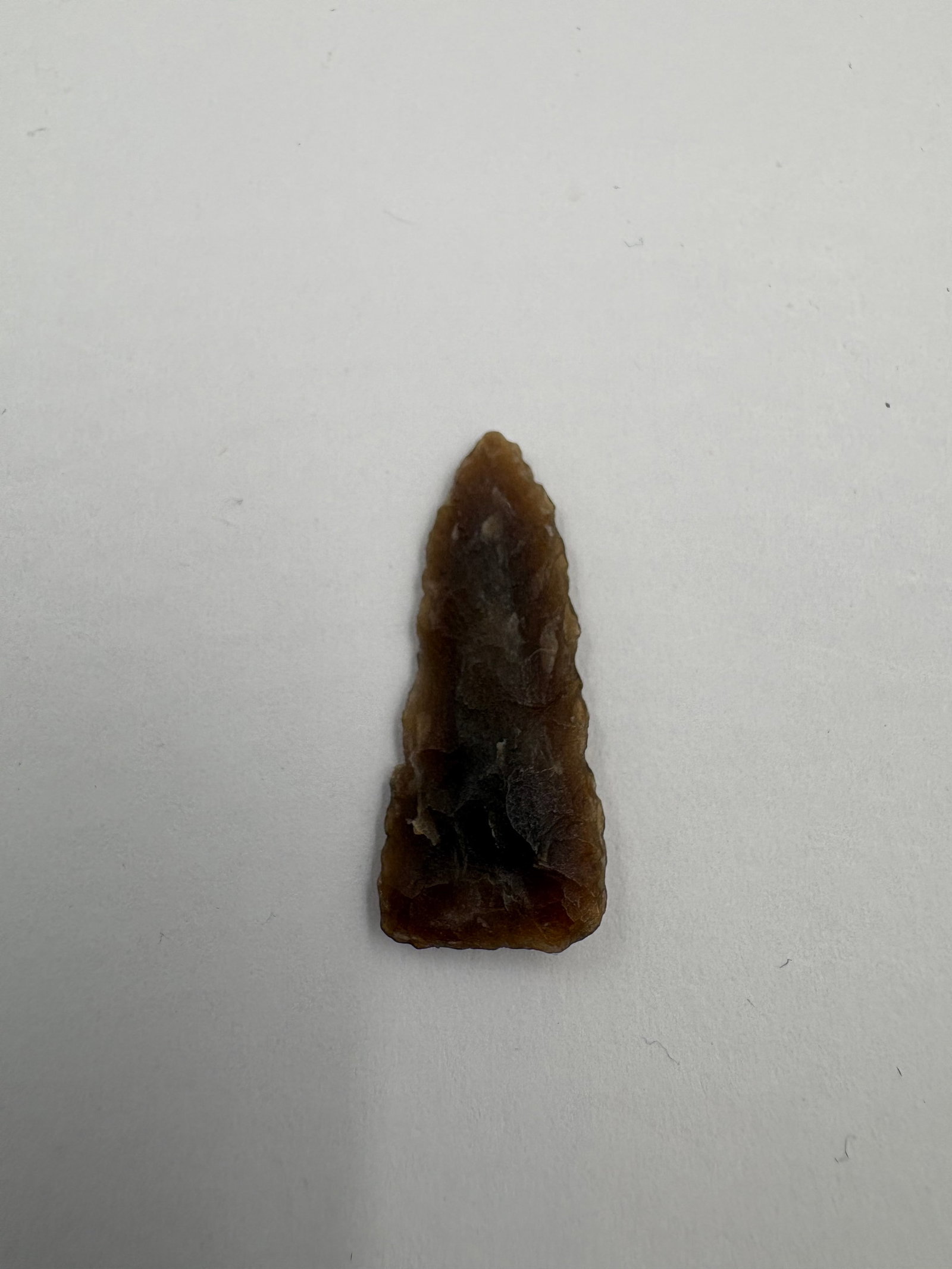 Plains Triangular Point, Motley COA: Triangular flint projectile point crafted from brown Knife River material, 1 1/16 inches long. Translucent amber-to-dark-brown coloration with a darker interior zone and visible flake scars on both
