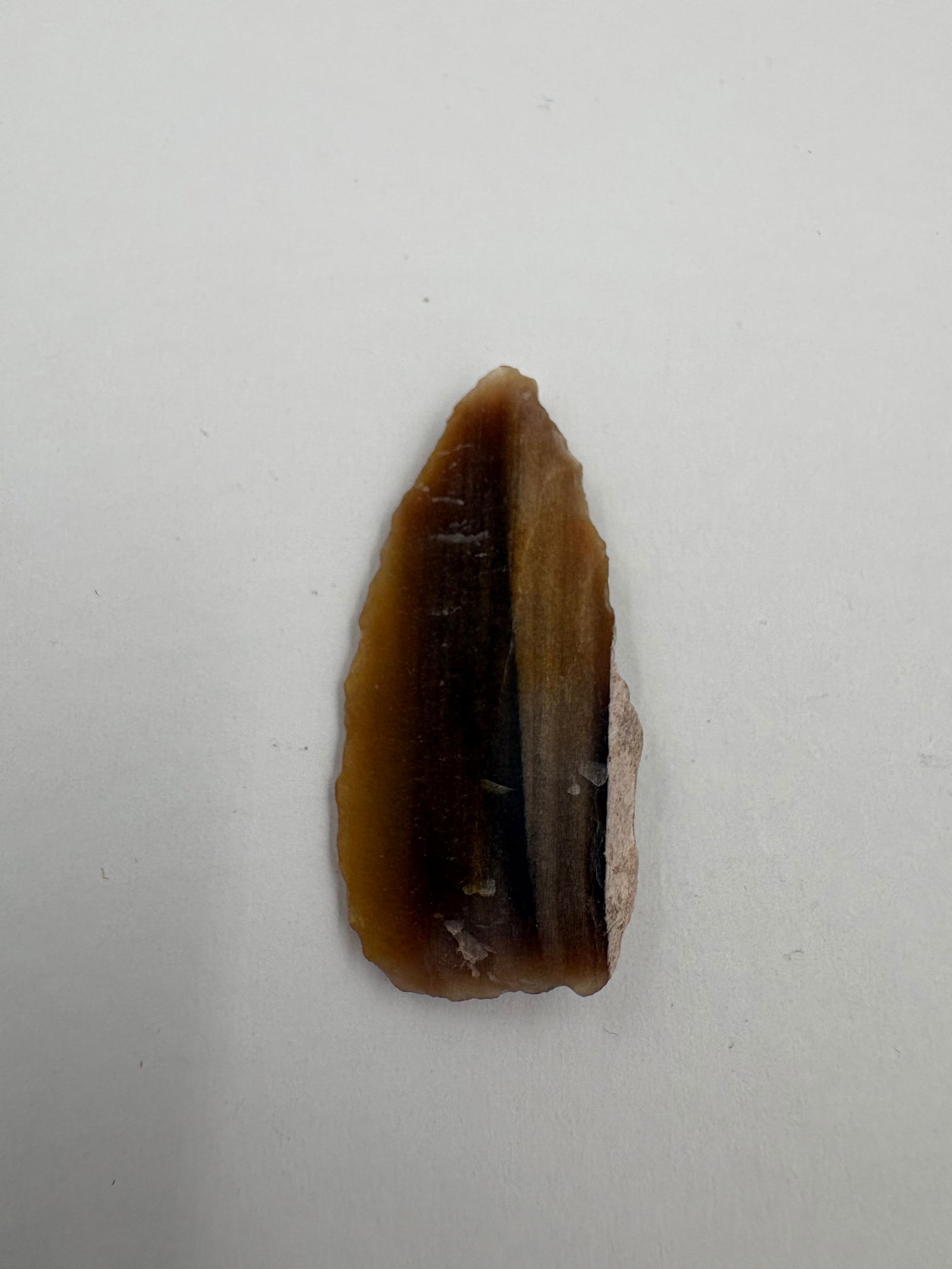 Western Agate Point: Translucent brown agate point with a tapered, leaf-shaped profile and distinct longitudinal banding. Both faces show regular flake scars and a narrow, sharp tip with a small remnant of lighter cortex