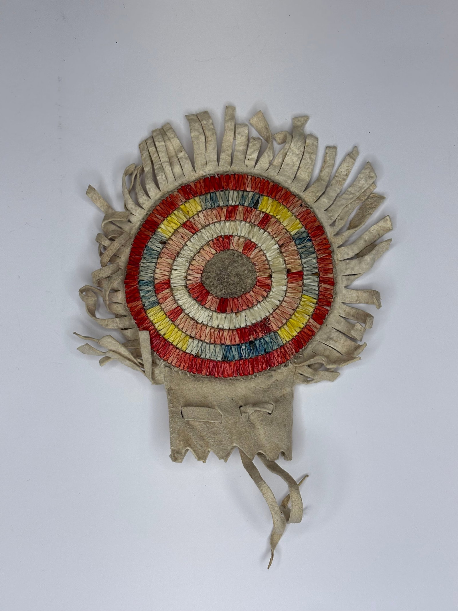 Quilled medallion pouch, turn of the century: Round quilled medallion pouch crafted from light tan leather with a fringed perimeter and concentric quillwork in red, yellow, blue, pink and cream. Center contains a circular felt insert surrounded