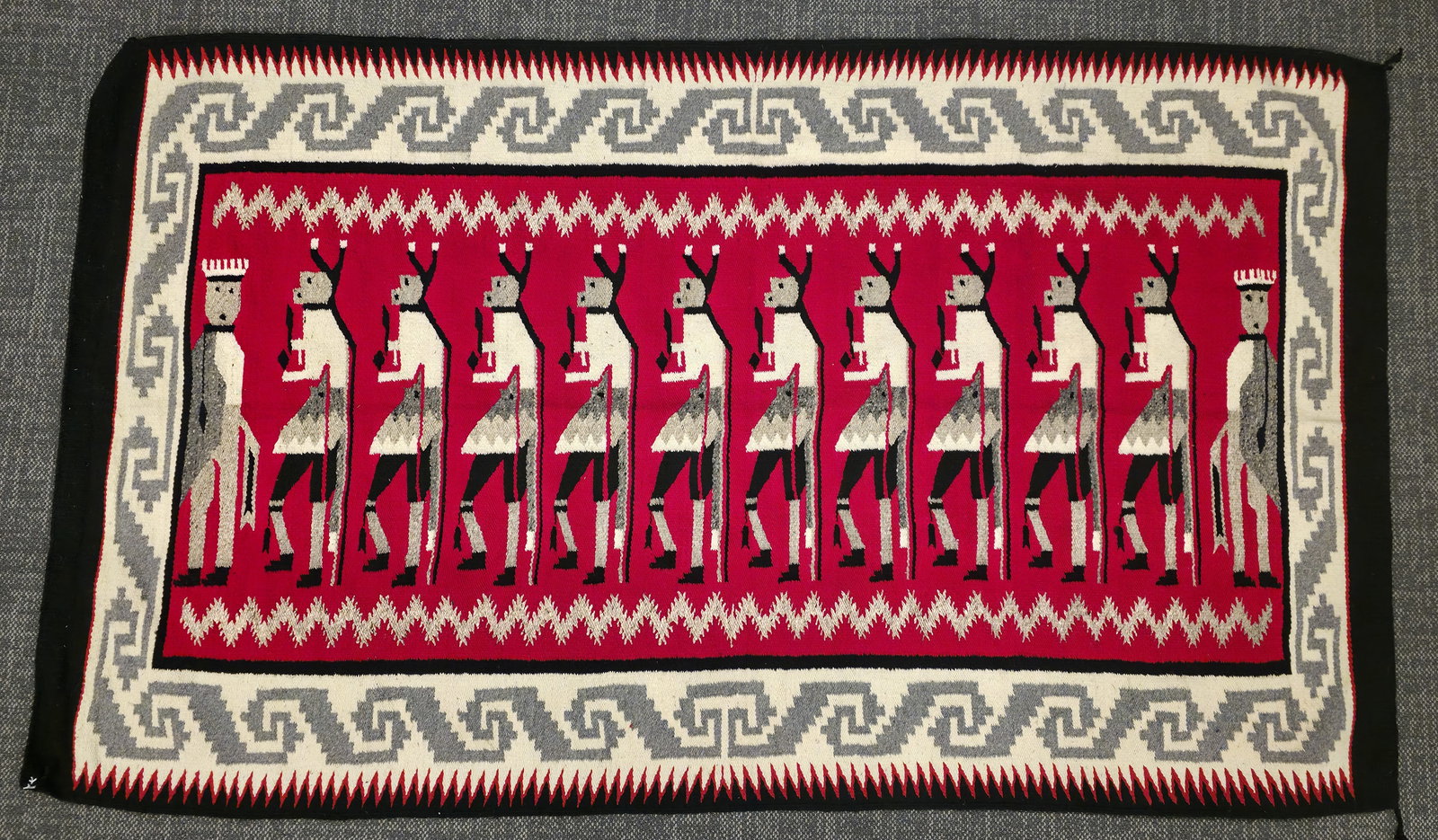 Navajo Weaving: Navajo-style weaving in a Yei design, featuring a central red field with a repeated row of stylized Yei figures worked in white, black and gray. Outer borders show stepped gray-and-white meander