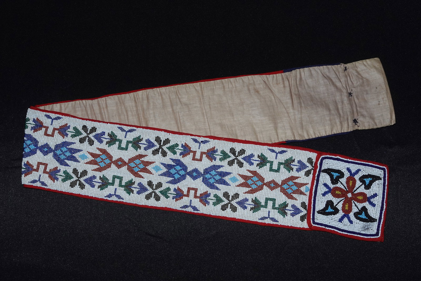 Loom Beaded Belt: Hand-loomed beaded belt attributed to the Cree, with a dense white seed-bead ground forming geometric floral and cross motifs in blue, green, red and black. Narrow red fabric binding frames the