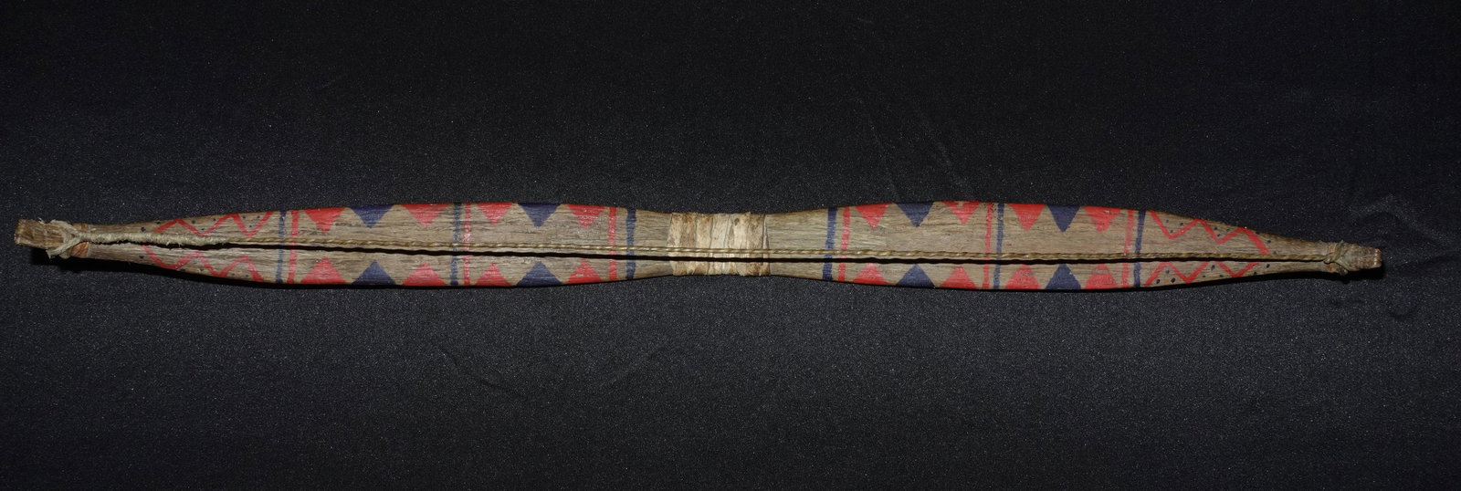 Cree Gauntlets: A wooden bow with a painted geometric motif of alternating red and blue triangles along one face and a smooth reddish-brown finish on the reverse. Twisted natural-fiber string is looped into notched