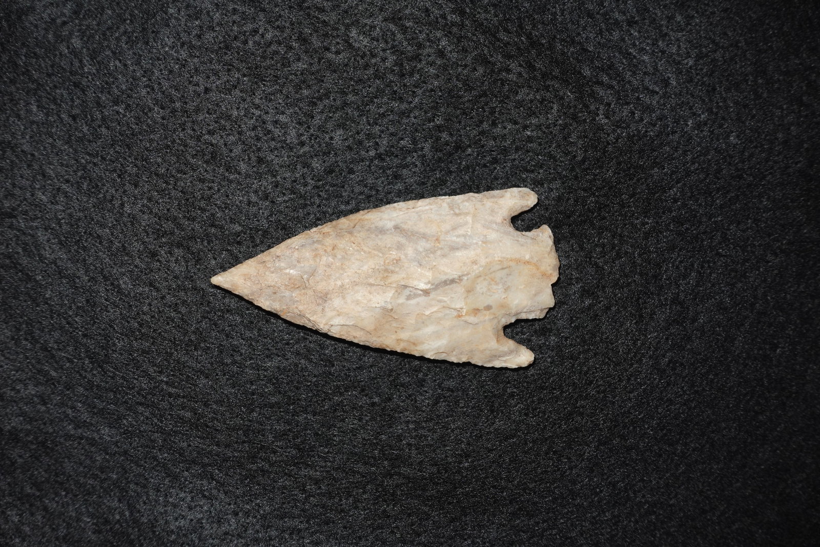 Smith: Light tan flaked stone projectile point with a narrow, elongated triangular blade and deep corner notches at the base. Bifacial flaking across both faces produces sharp, leveled edges and visible