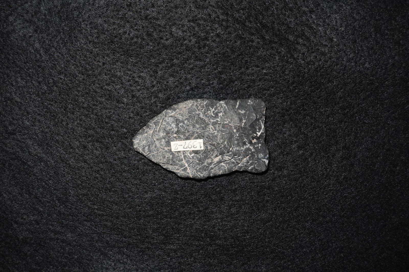 Quad: Dark gray to black angular stone with pale white, vein-like streaks across the surface. Matte, fine-grained texture with visible fracture lines and irregular, flaked edges. Broken margins display