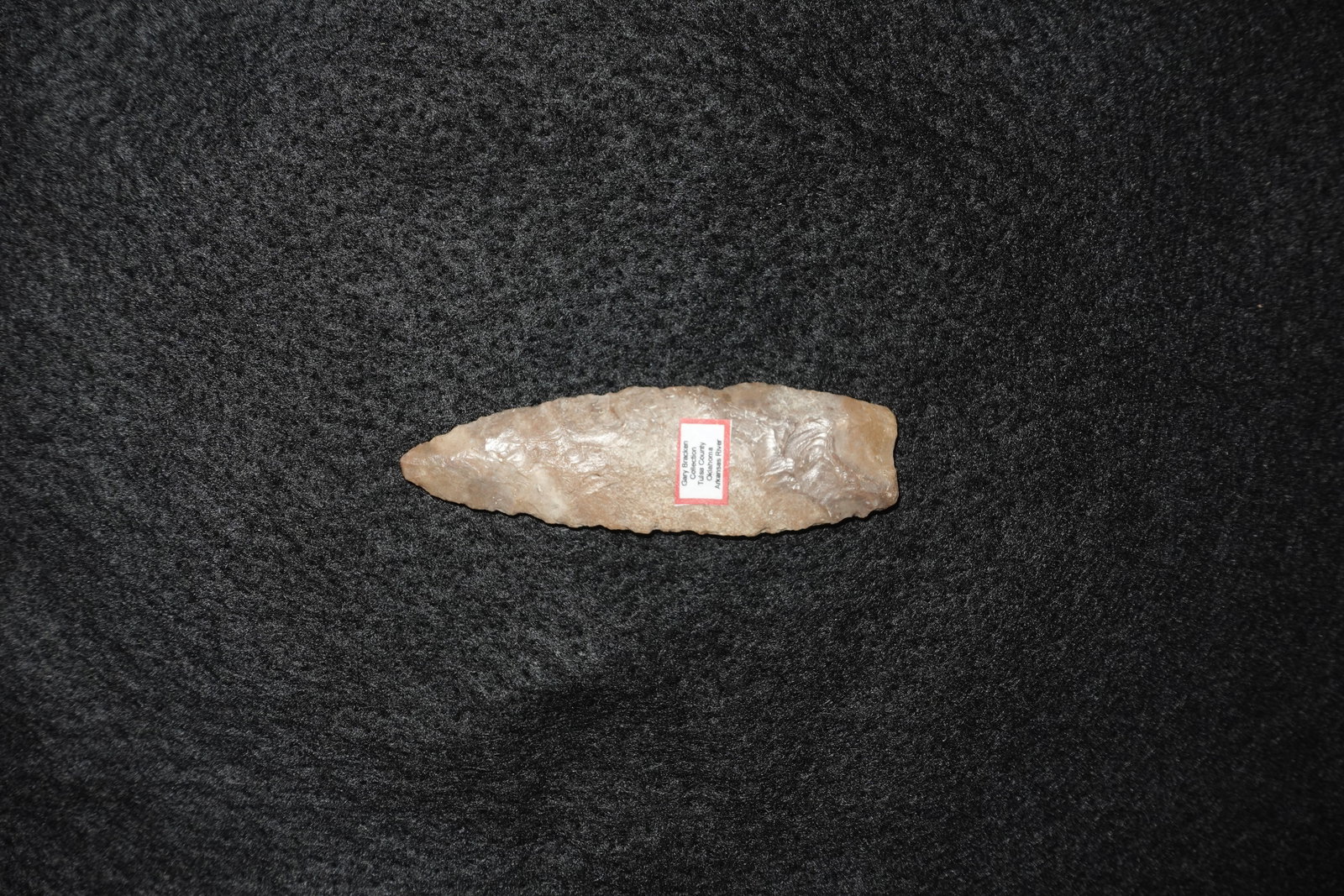 Midland: Elongated lanceolate flaked stone projectile point with tan-beige translucent coloring and bifacial flake scars across both faces. Edges show careful pressure flaking producing subtle serrations