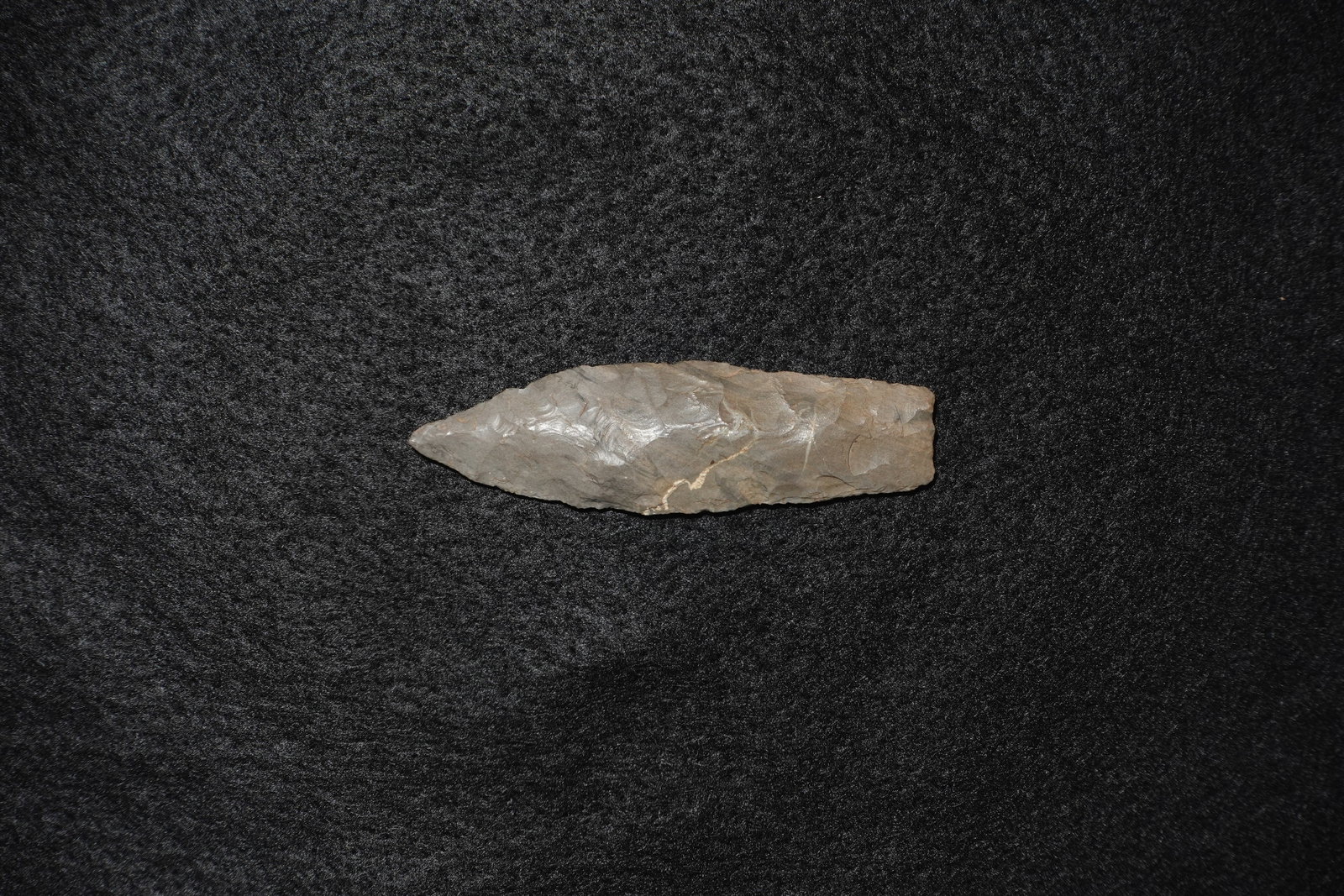 Agate Basin: Long narrow flaked stone projectile point with bifacial thinning and a tapered tip. Gray-brown chert shows overlapping flake scars and a faint light-colored mineral vein on one face. Edges display fin