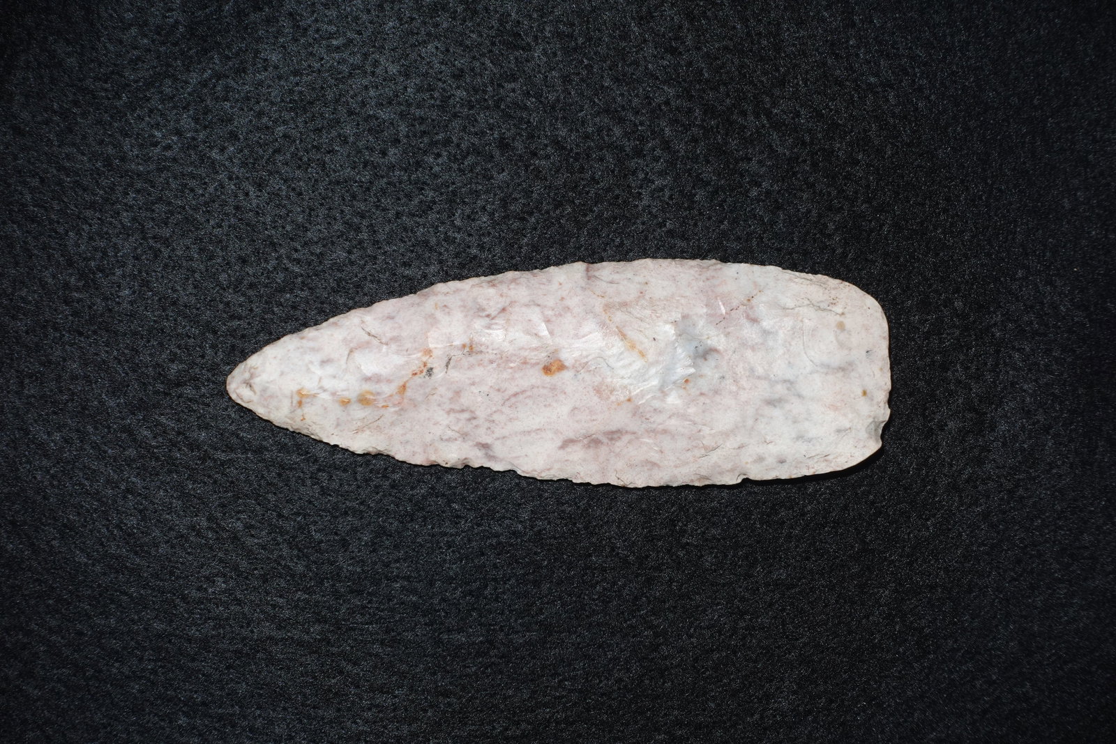 Sedalia: Leaf-shaped bifacial stone point with pale pink to off-white coloration and subtle brown-orange staining along the central face. Narrow, tapered profile with fine pressure flaking along the edges and