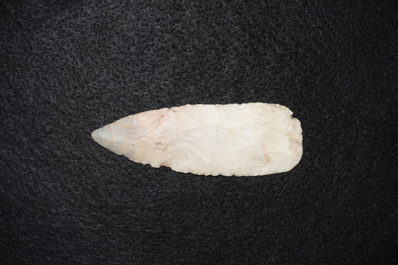 Paleo Knife: Elongated leaf-shaped flaked stone blade in off-white to cream tones. Bifacial flaking covers both faces with percussion and pressure flake scars; edges display fine retouching and a subtly serrated l
