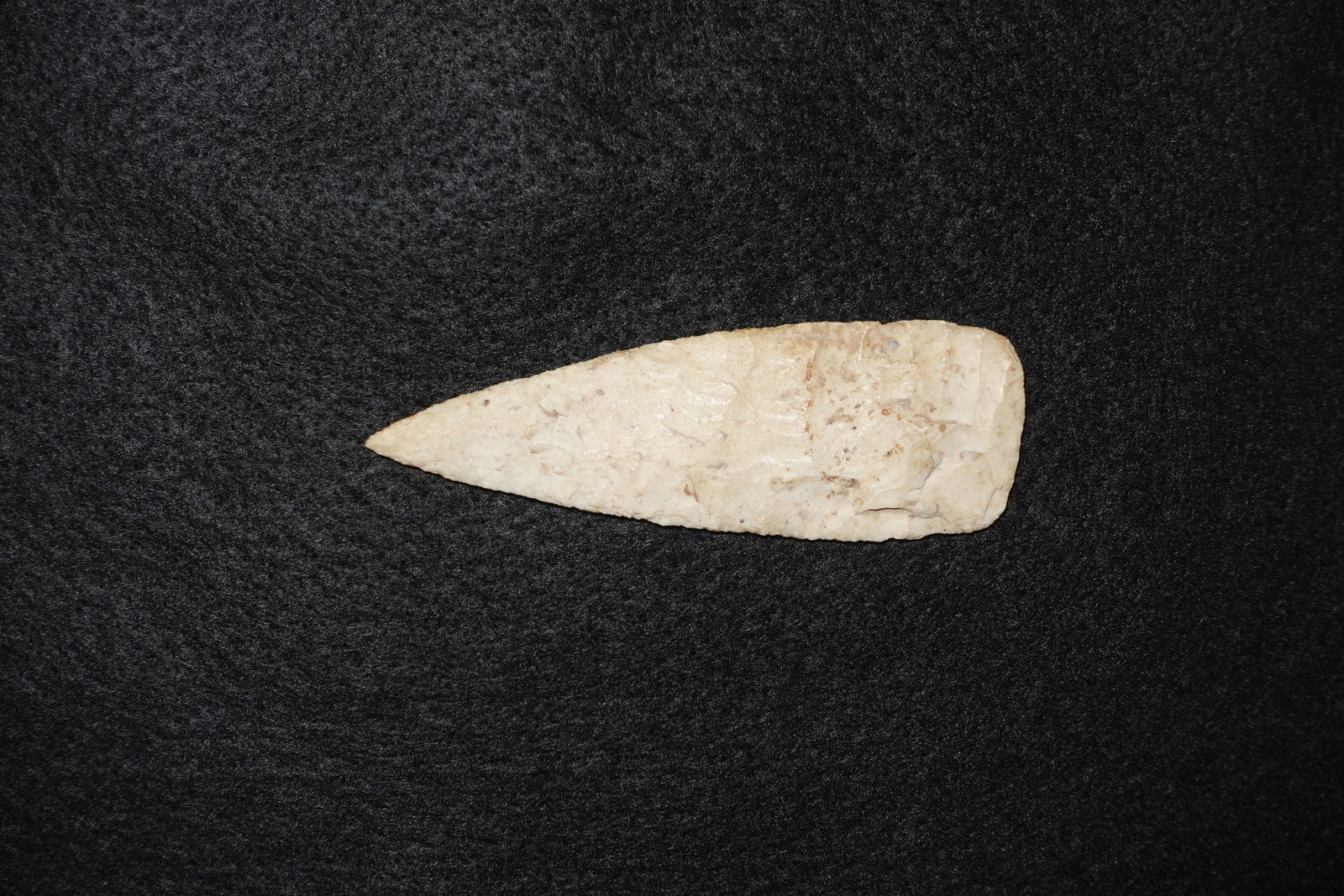 Fine Paleo Blade: Pale cream-to-tan flaked stone blade with a narrow, pointed tip and a rounded, smoothed base. Both faces exhibit stepped flake scars and fine bifacial working along the edges, creating a thin,