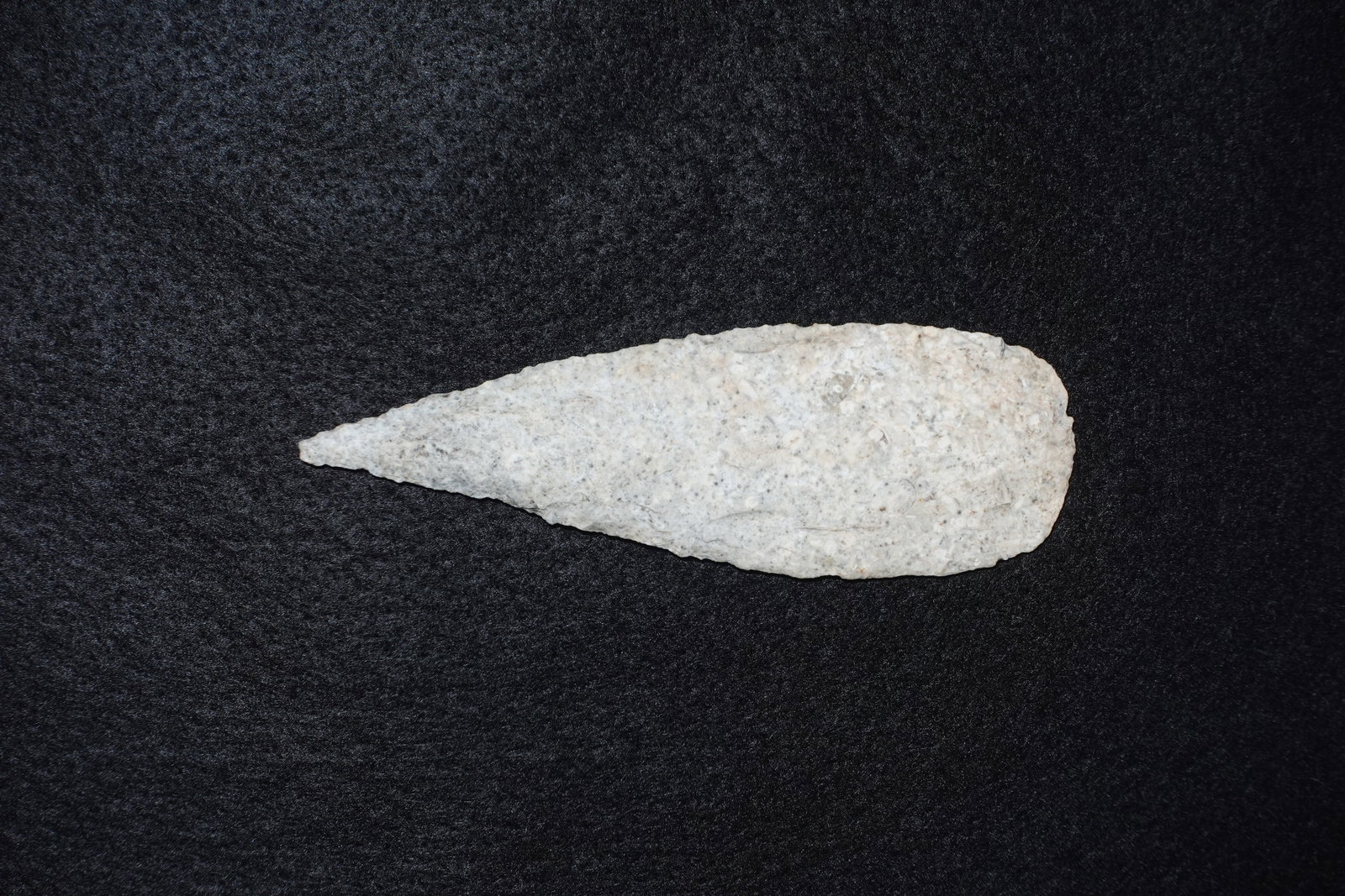 Cobbs: Elongated lanceolate stone projectile point with a narrow tapered tip and a broad rounded base. Light gray to off-white material shows dense flaking scars and fine bifacial retouch along the edges,