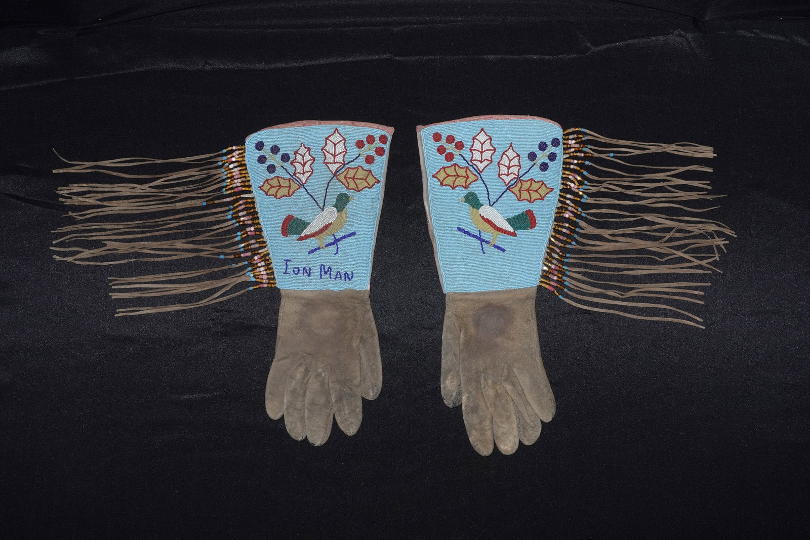 Plains beaded gauntlets: Beaded leather gauntlets with sky-blue seed-bead cuffs depicting a perched bird surrounded by stylized leaves and berry clusters in red, white, yellow and green. Pink trim borders the cuff tops and