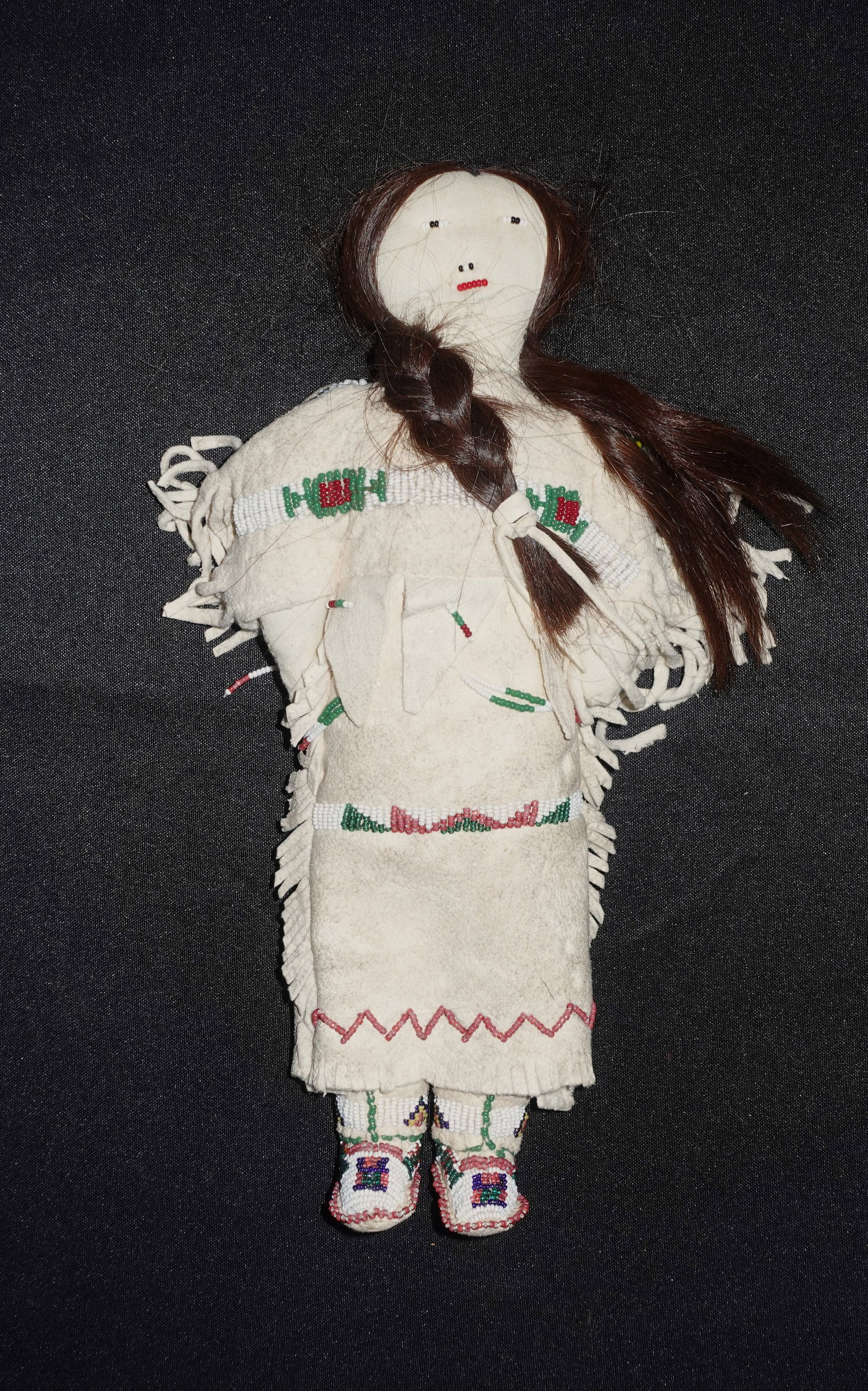 Sioux Doll: Handmade cloth doll wearing a cream-colored soft leather dress trimmed with fringe and bands of white seed beads with red and green geometric motifs. Long brown hair is braided and tied at the front,