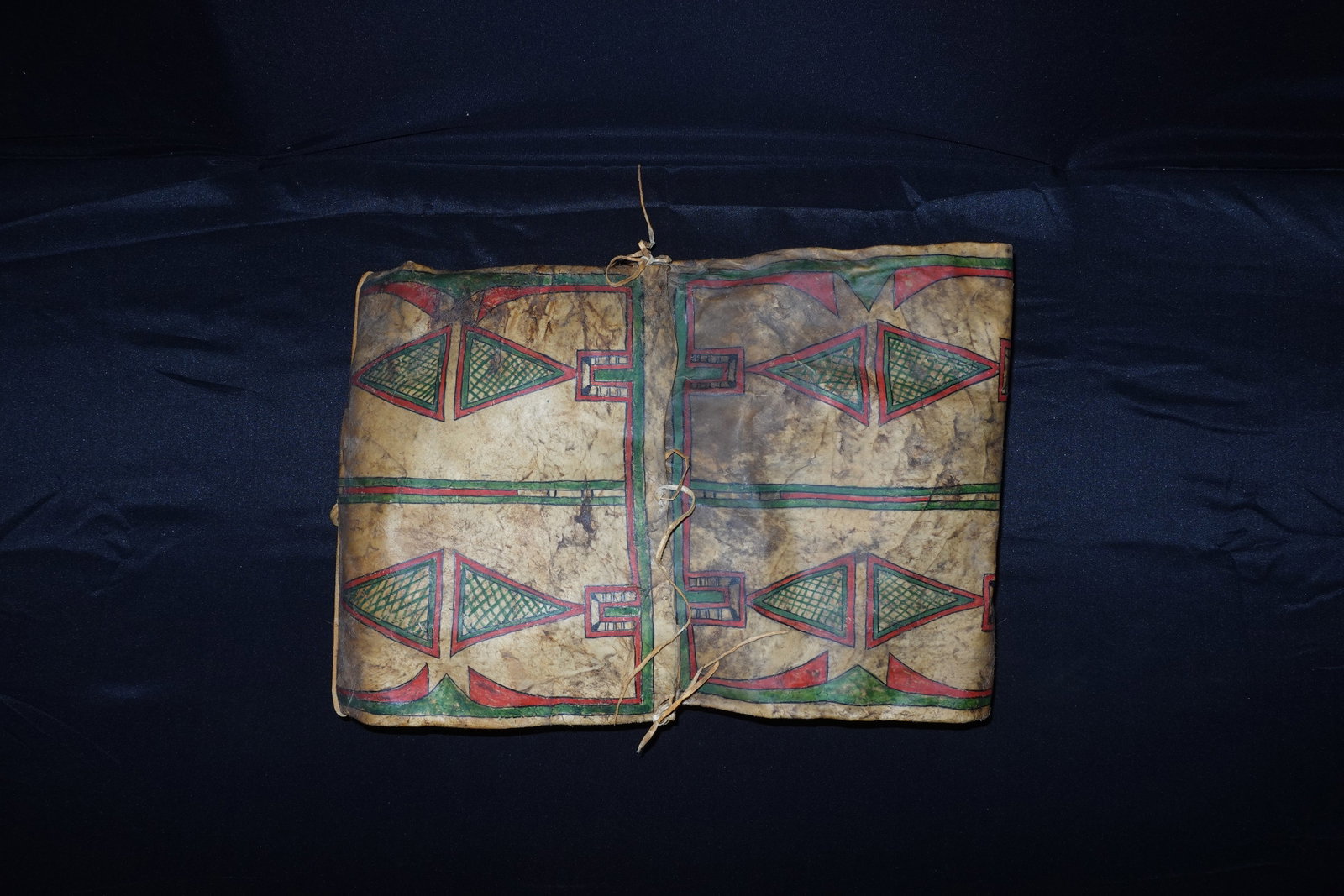 Northern Plains Parfleche: Rawhide parfleche with hand-painted geometric motifs in red, green and black framed by parallel border bands. Folded panels fasten with leather thongs and edge lacing, forming a tied closure down the