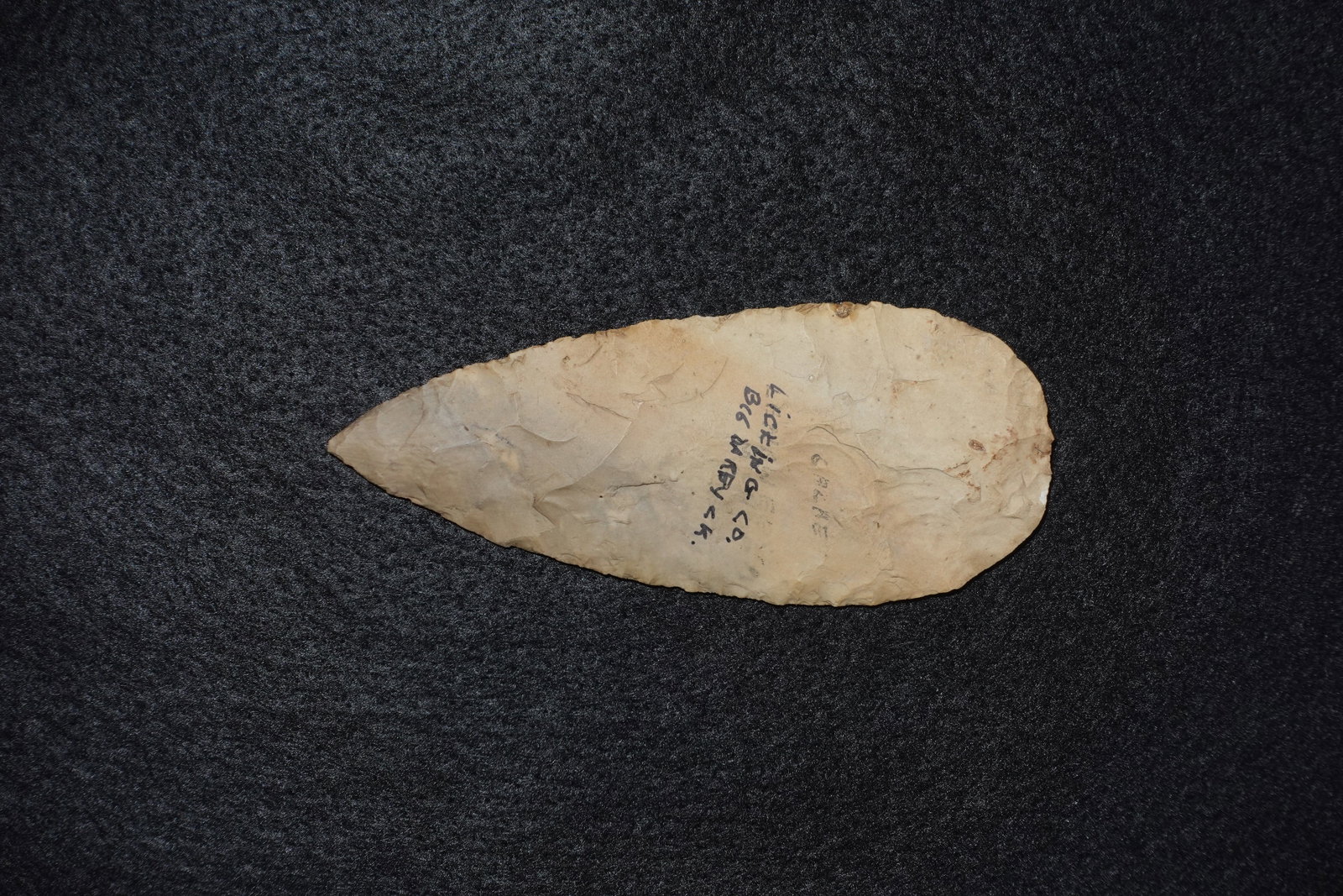 Cache Blade #1: Flaked stone cache blade in pale tan-to-cream chert with bifacial flaking and a tapered, pointed profile. One face has black ink handwriting noting Licking Co., and the surface shows natural mineral