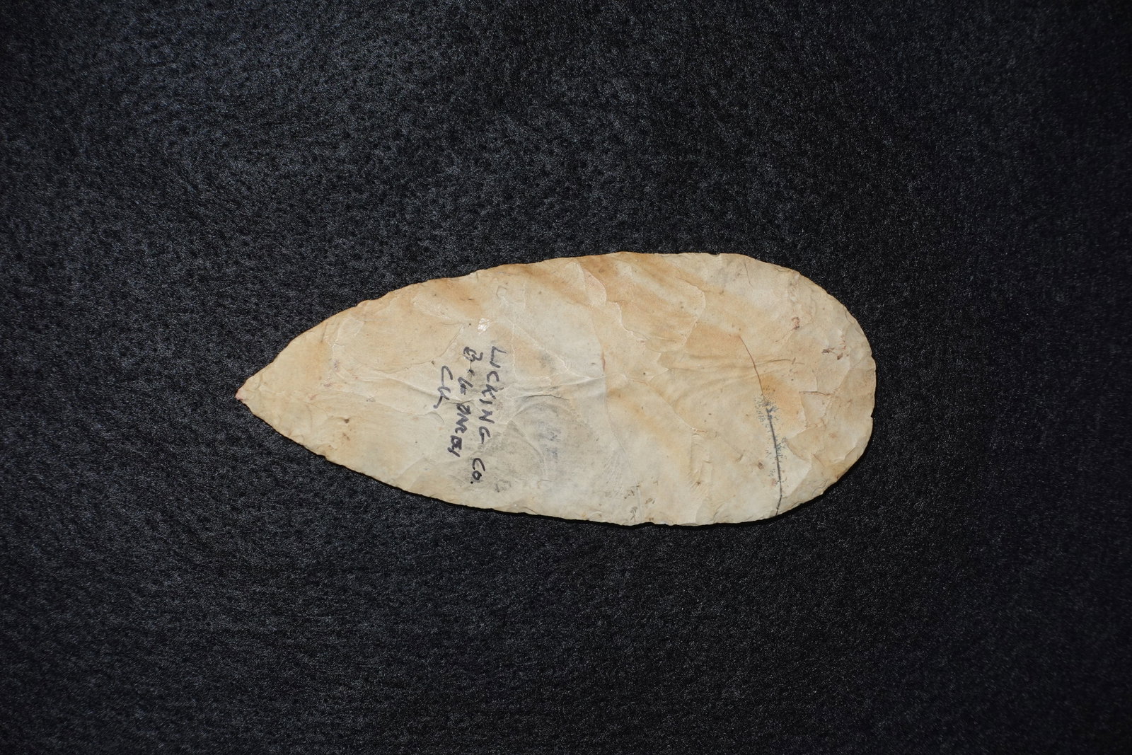 Cache Blade #2: Light tan hand-flaked stone blade with an elongated oval-knife profile and a slightly pointed tip. Bifacial flaking produces visible knapped flake scars across both faces and a straight to slightly