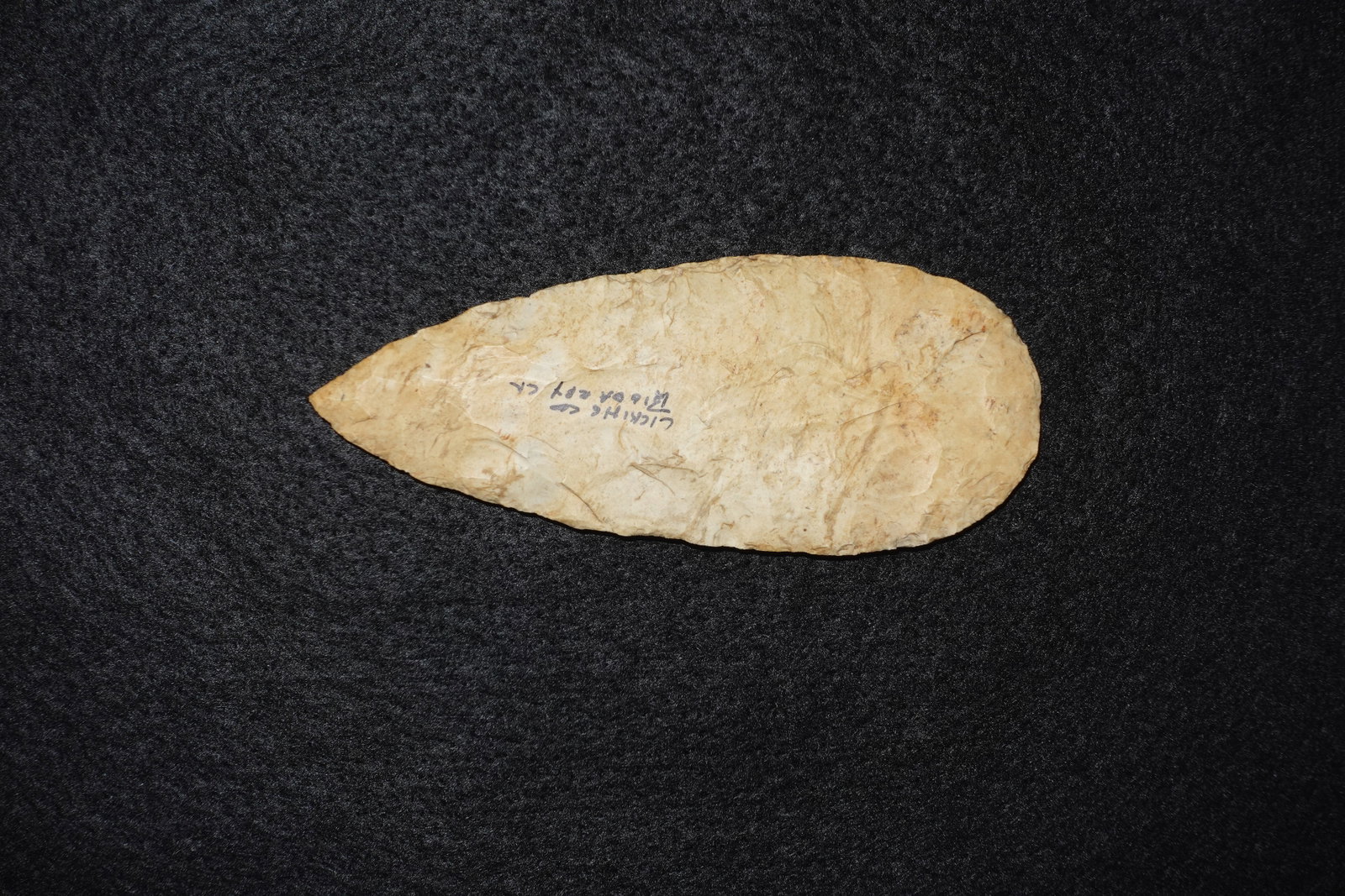 Cache Blade #4: Leaf-shaped flaked stone blade with a pointed tip and broad rounded base, worked bifacially along the edges. Light tan to creamy color with visible flake scars and subtle mineral banding across the