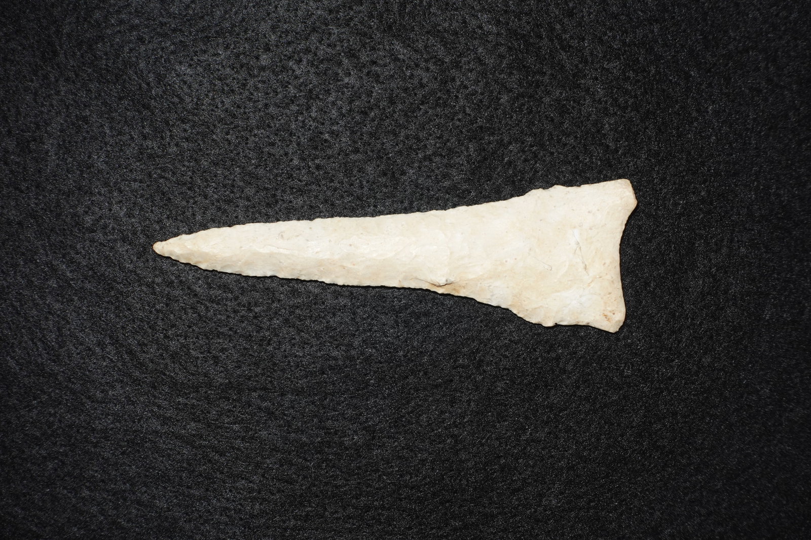Dalton: Long, narrow flaked-stone projectile point with a sharply tapered tip and a broad triangular basal stem. Light tan to cream surface with visible pressure-flake scars and fine marginal retouch along