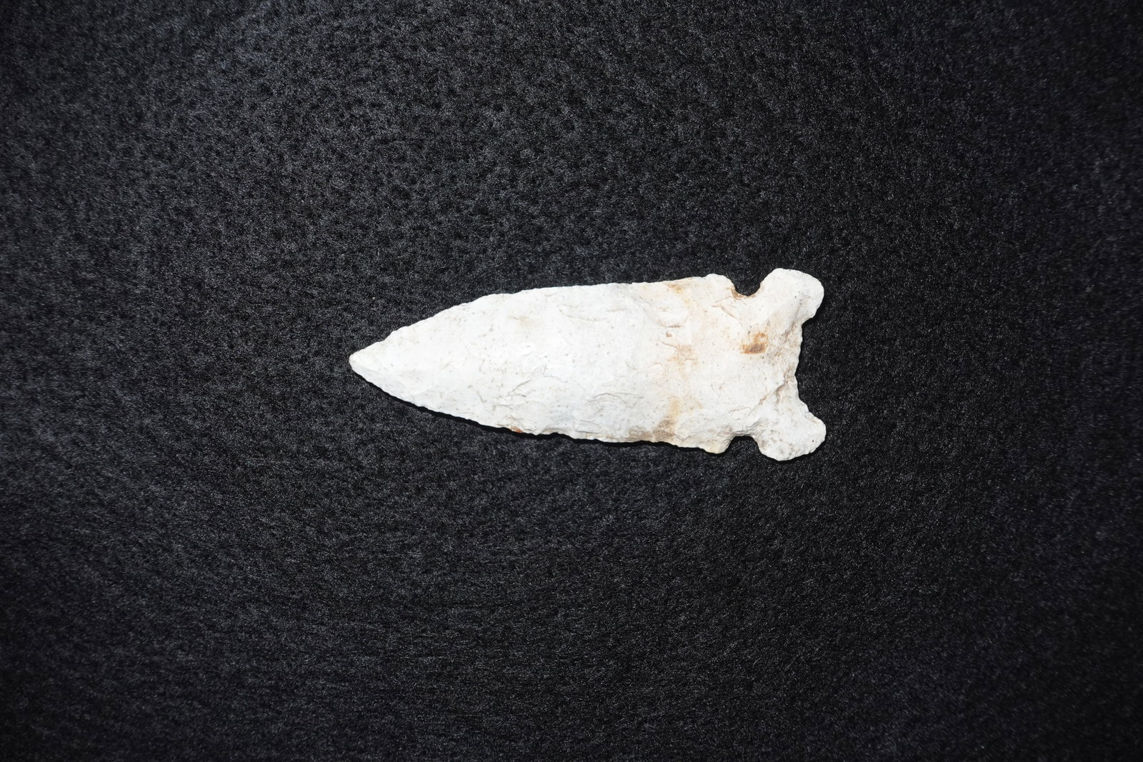 Graham Cave: Light-colored stone projectile point with a narrow lanceolate blade and a stemmed, slightly notched base. Bifacial flake scars and fine edge retouch are visible across both faces, with areas of tan