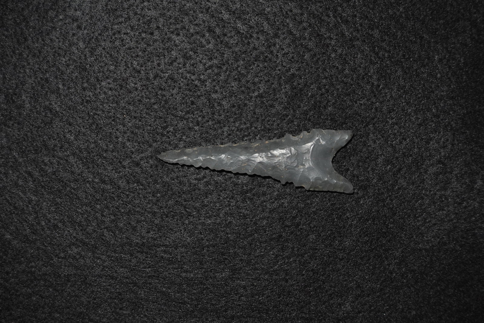 Dalton: Dark gray to black knappable stone projectile point with deep basal notches and a pronounced stem. Bifacial flaking produces overlapping flake scars and a glossy finish consistent with obsidian or