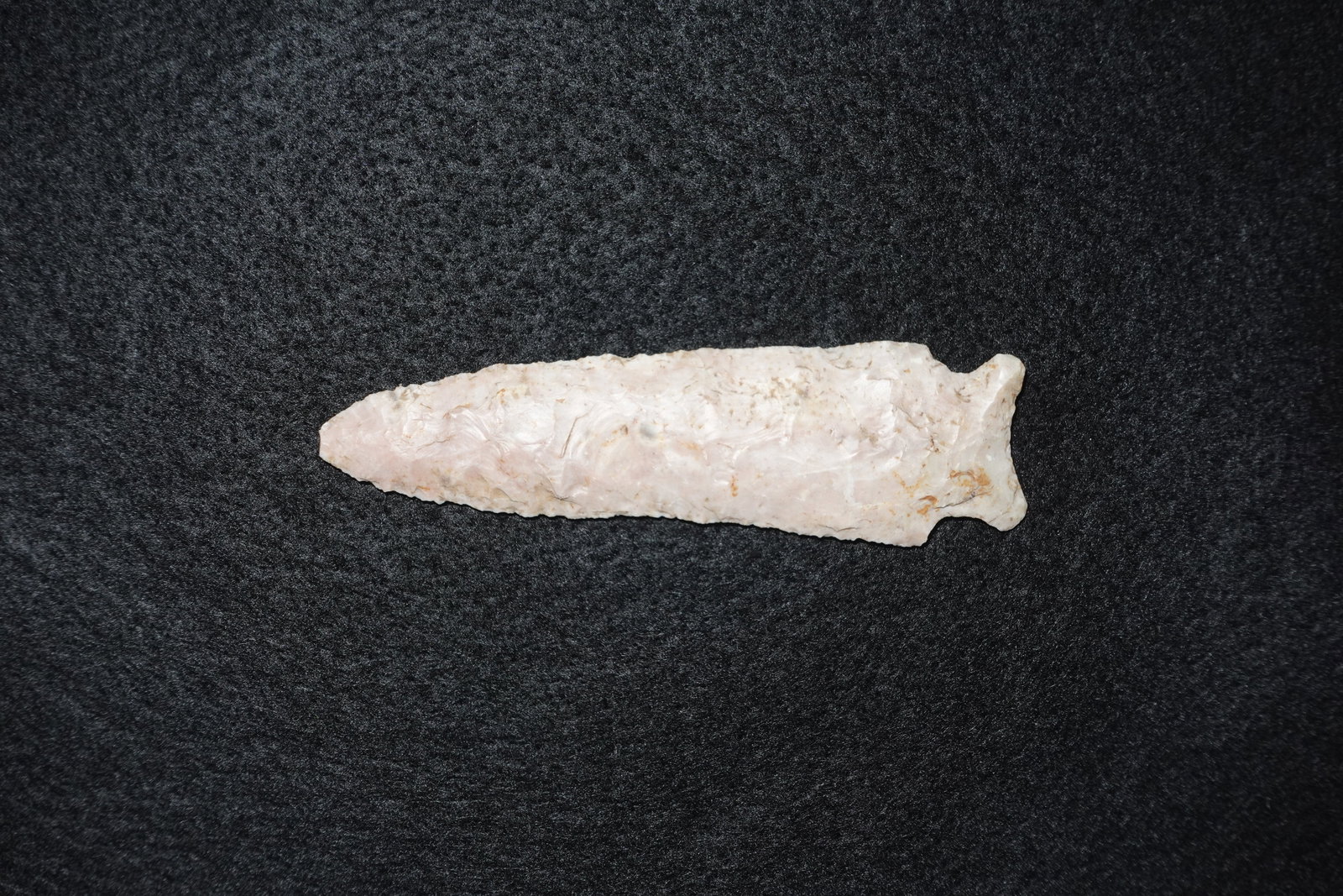 Stillwell: Elongated bifacial stone projectile point with a tapered blade and a distinct notched stem at the base. Crafted from a pale cream to light pink translucent stone with prominent flake scars and fine