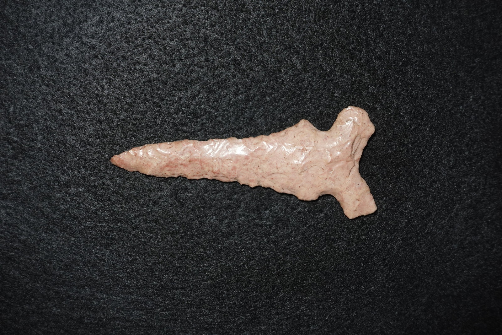 Graham Cave: Pinkish-beige flaked stone projectile point with a long, narrow triangular blade and distinct basal notches forming short ears. Bifacial flaking and fine edge retouch are apparent along the blade;
