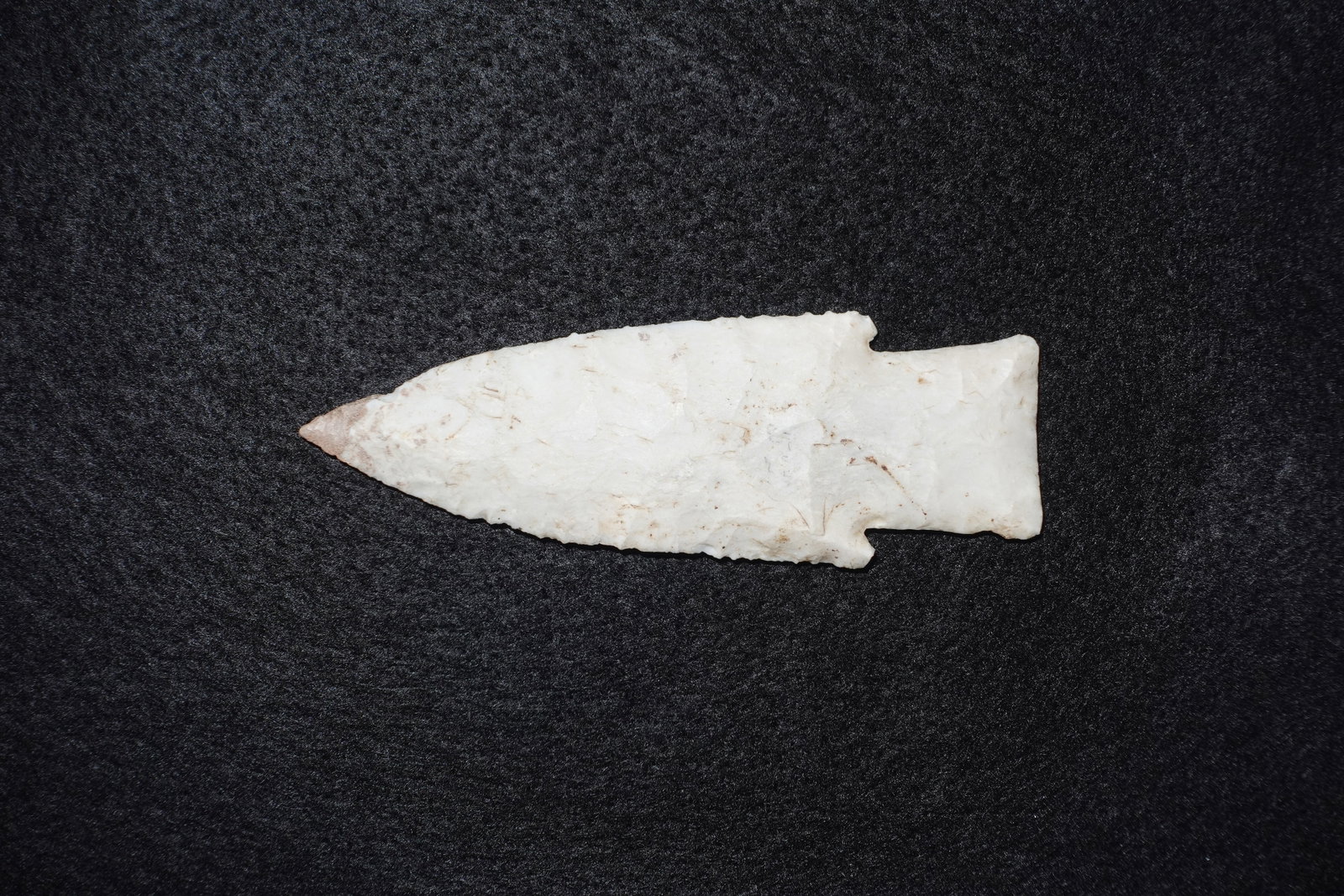 Hardin: White flaked stone projectile point with a lanceolate blade, serrated edges, and a narrow stem terminating in a notched, bifurcate-style base. Smooth translucent white surface shows light tan staining