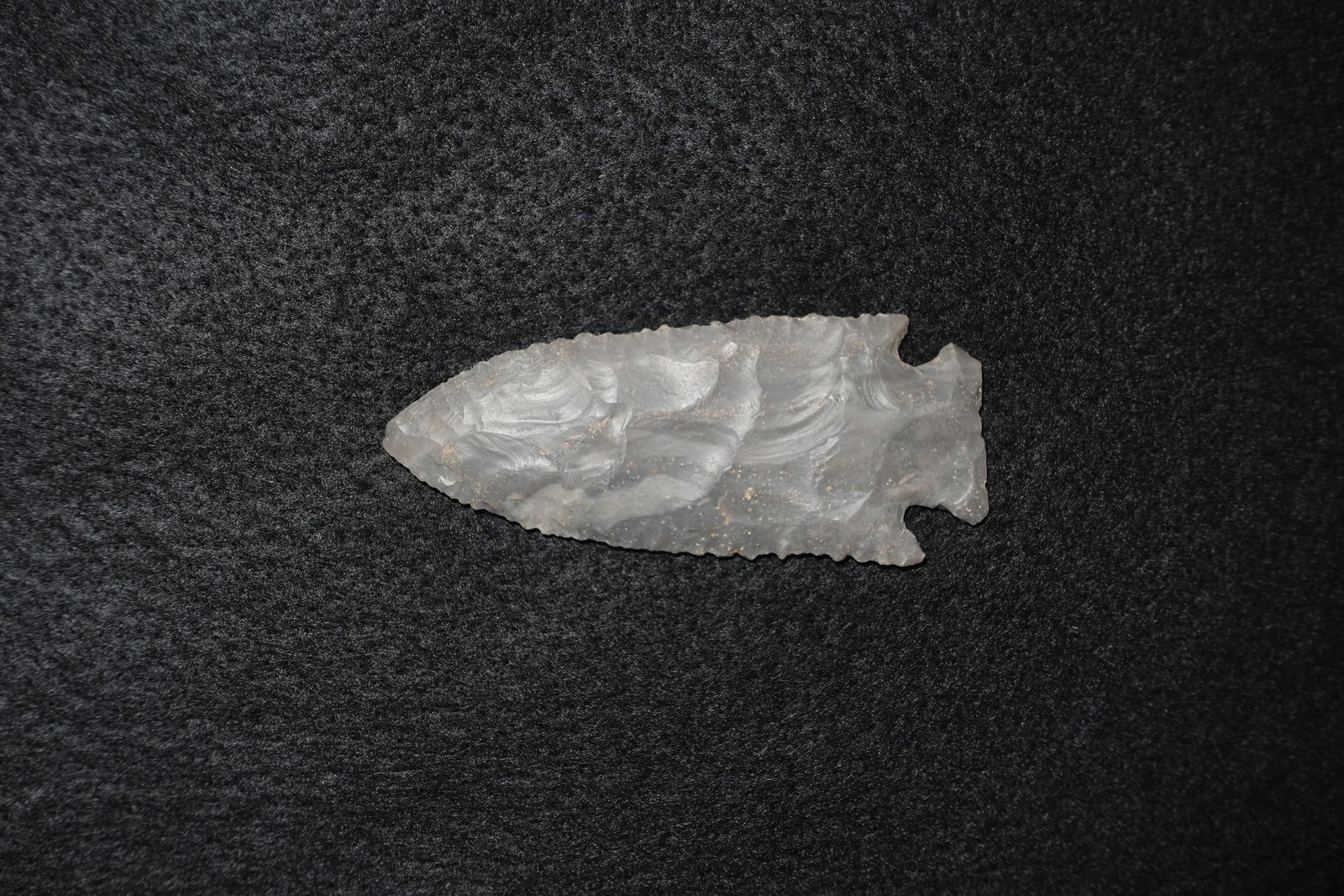 Hardin: Translucent gray stone projectile point with an elongated leaf-shaped blade and finely flaked, serrated edges. Bifacial flaking produces pronounced ripple-like flake scars across both faces, and a