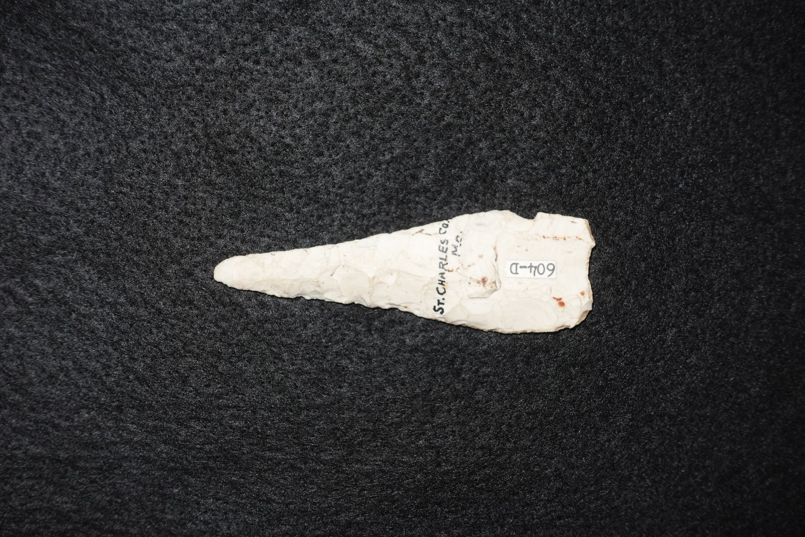 Clovis: Cream-colored flaked stone projectile point with a long, narrow tapering blade and a broader rounded base. Both faces show bifacial flake removals and fine edge flaking, with light surface staining