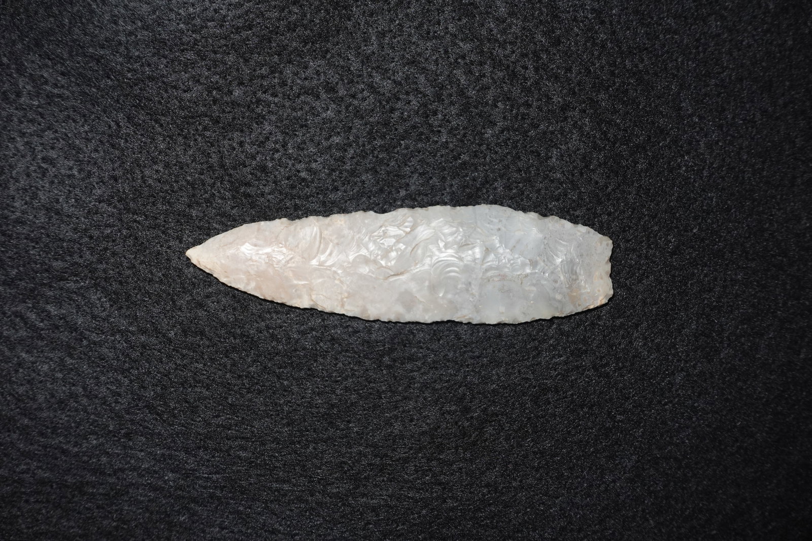 Angostura: Elongated lanceolate flaked stone point in a translucent pale-white tone with minor tan mottling. Both faces display dense bifacial pressure flaking with pronounced feathered flake scars and a sharp